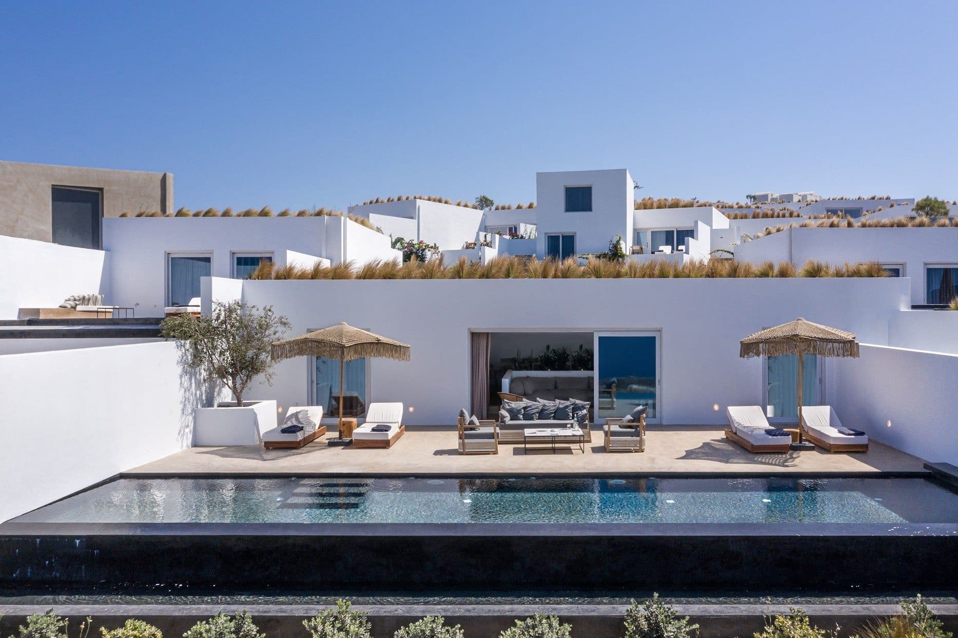 Infinity pool waters mirror azure skies while natural fiber umbrellas cast gentle shadows over pristine loungers at this whitewashed Cycladic retreat.
