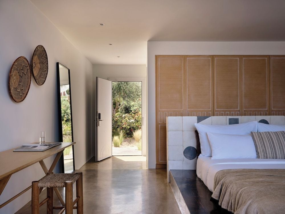 Natural light floods through French doors into this serene suite, where woven baskets and warm wood create an understated Mediterranean elegance that feels both luxurious and grounding.