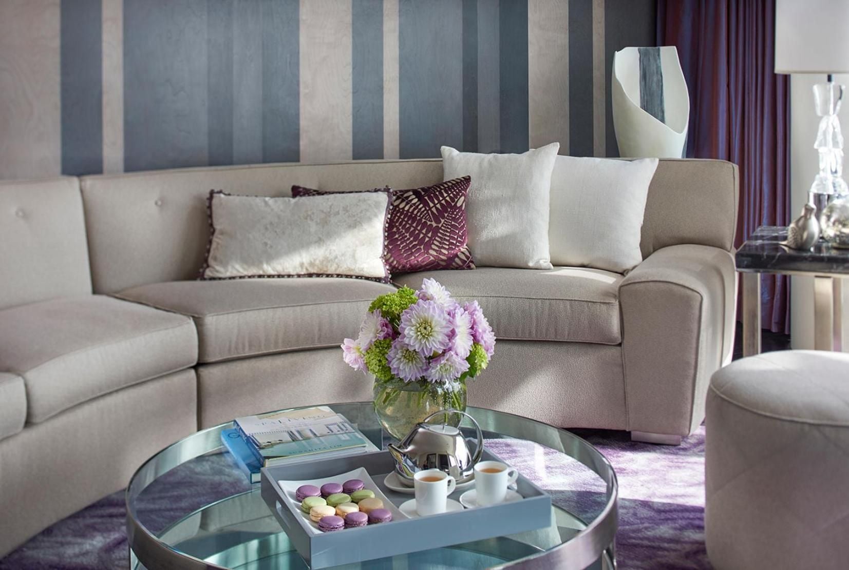 Plush sectional seating and glass coffee table create an intimate gathering space, where purple blooms and macarons await in this elegantly appointed suite living area.