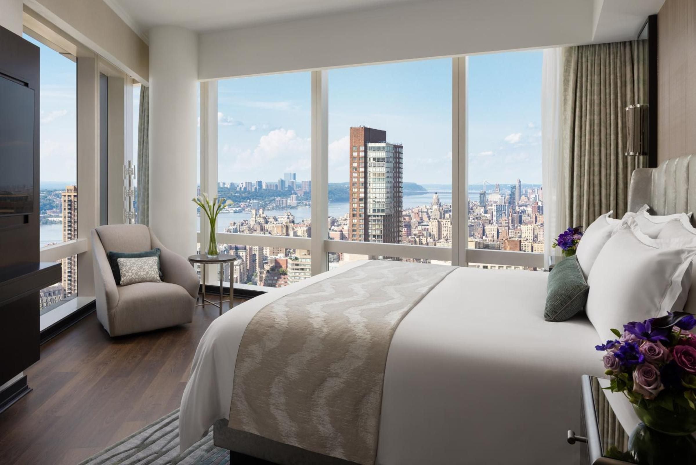 Floor-to-ceiling windows frame Manhattan's skyline from this serene sanctuary, where crisp linens meet hardwood floors and purple blooms add warmth to contemporary elegance.