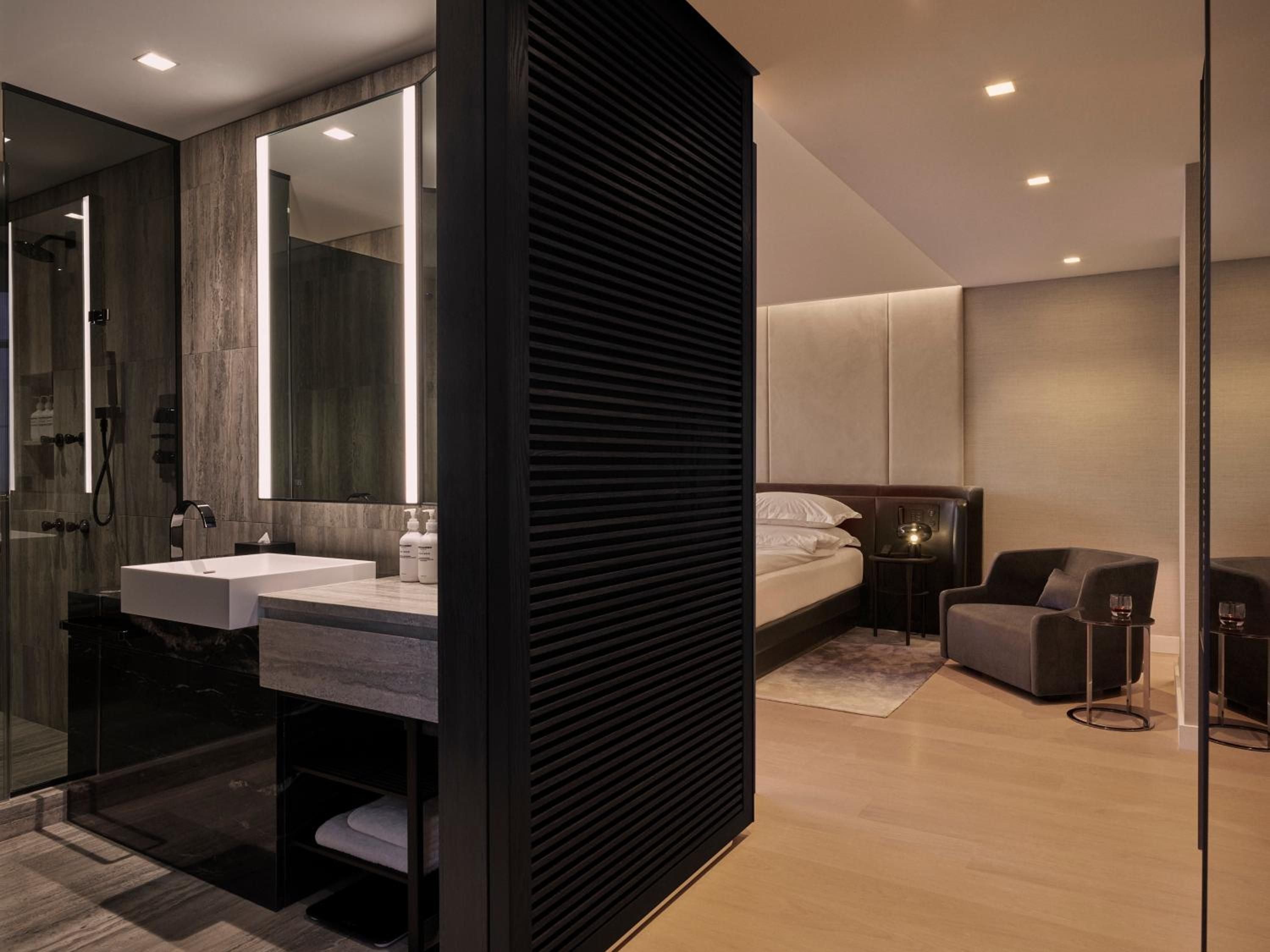 Floor-to-ceiling glass and sleek black slat dividers create seamless flow between marble-clad bath and minimalist sleeping quarters, where warm lighting softens geometric lines.