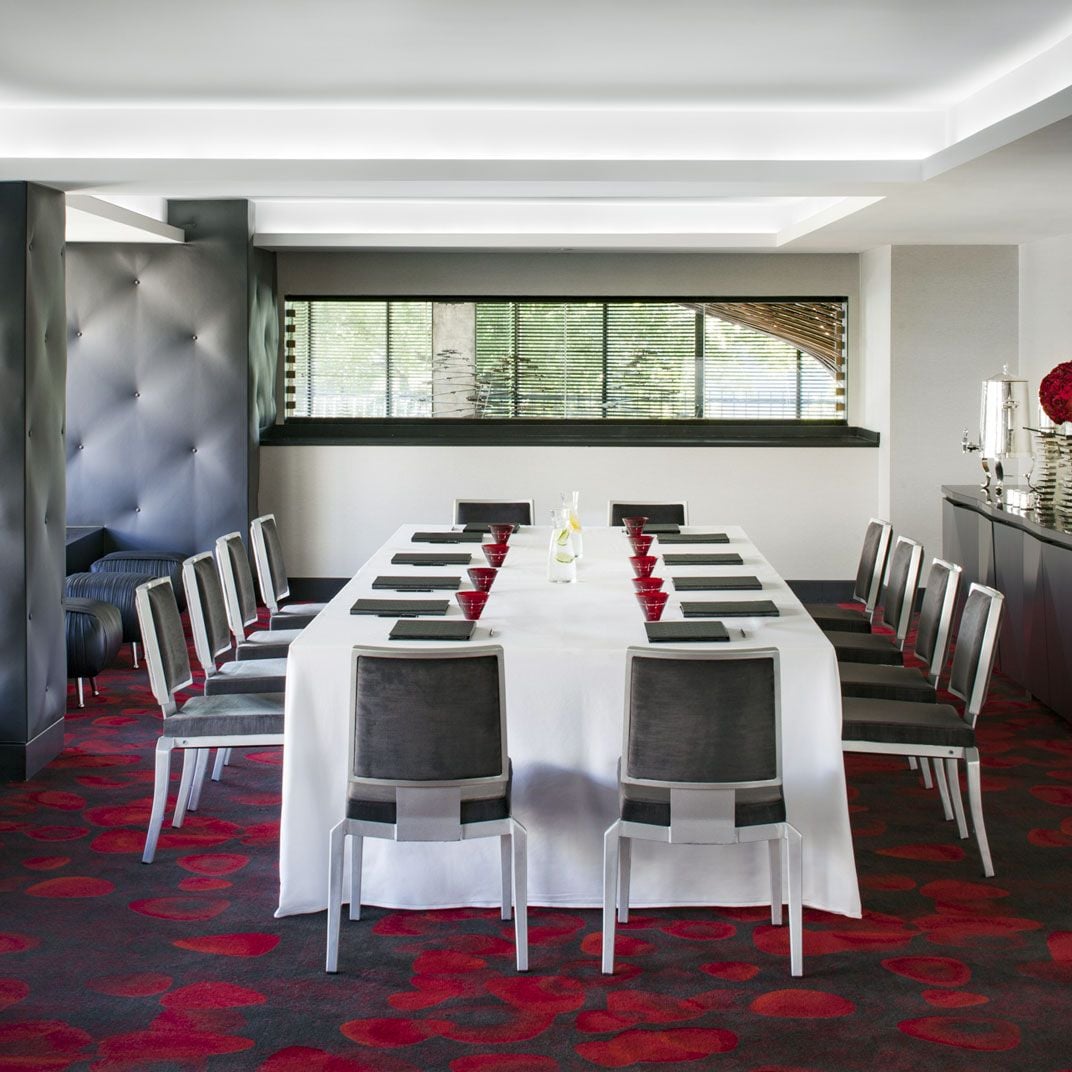 Crimson-patterned carpeting and sleek gray chairs create an intimate boardroom where power lunches unfold beneath dramatic tufted banquettes and floor-to-ceiling windows.
