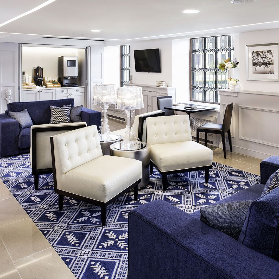 Navy geometric rugs anchor cream leather chairs in this sophisticated suite, where nautical blues meet crisp whites and natural light streams through patterned window treatments.