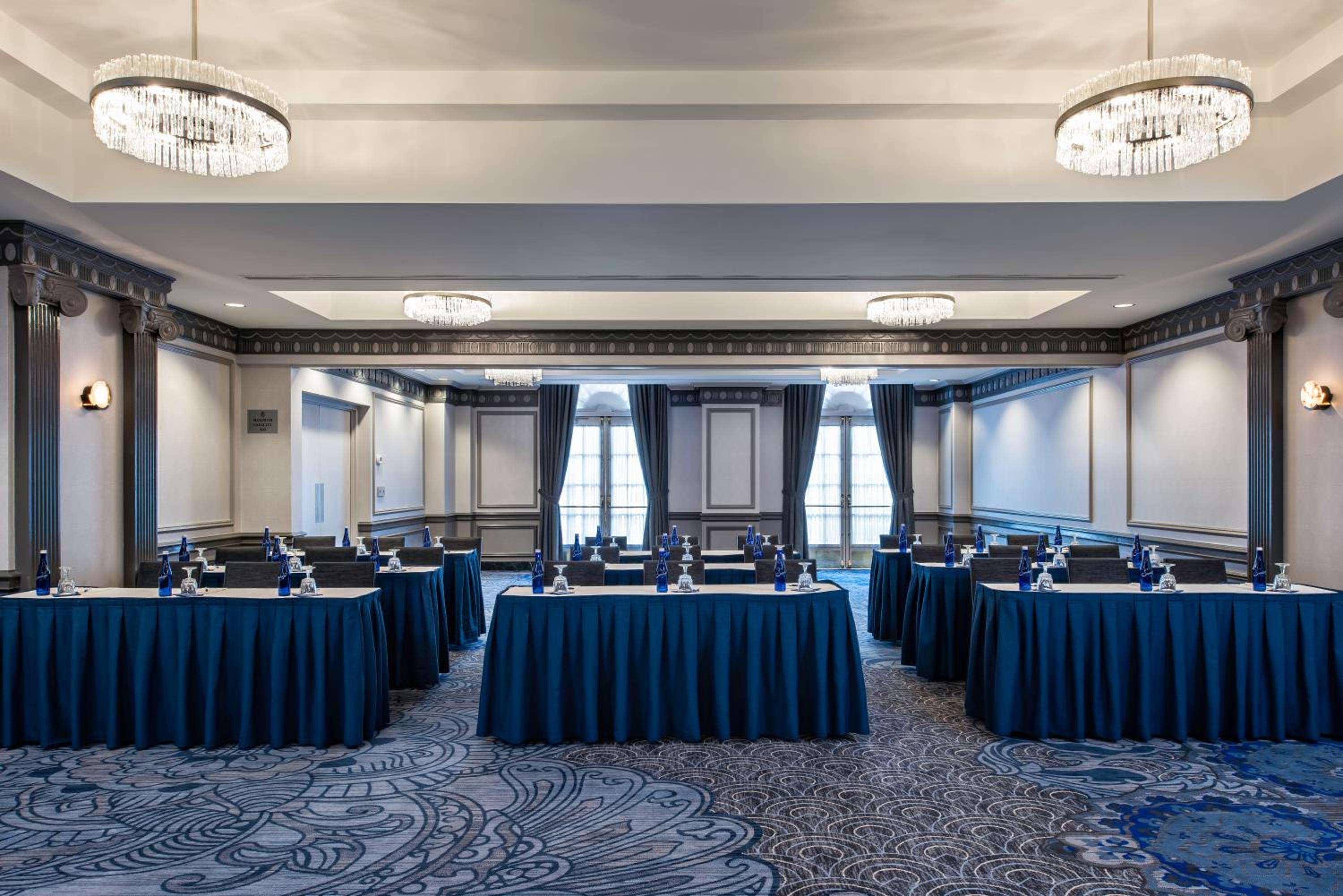 Crystal chandeliers cast warm light over navy-draped tables in this stately meeting space, where ornate columns and coffered ceilings create an atmosphere of refined gravitas.
