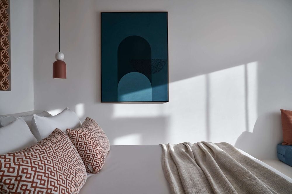 Geometric terracotta pillows and abstract art create sophisticated contrast against crisp white linens, while natural sunlight streams across minimalist surfaces in this serene suite.