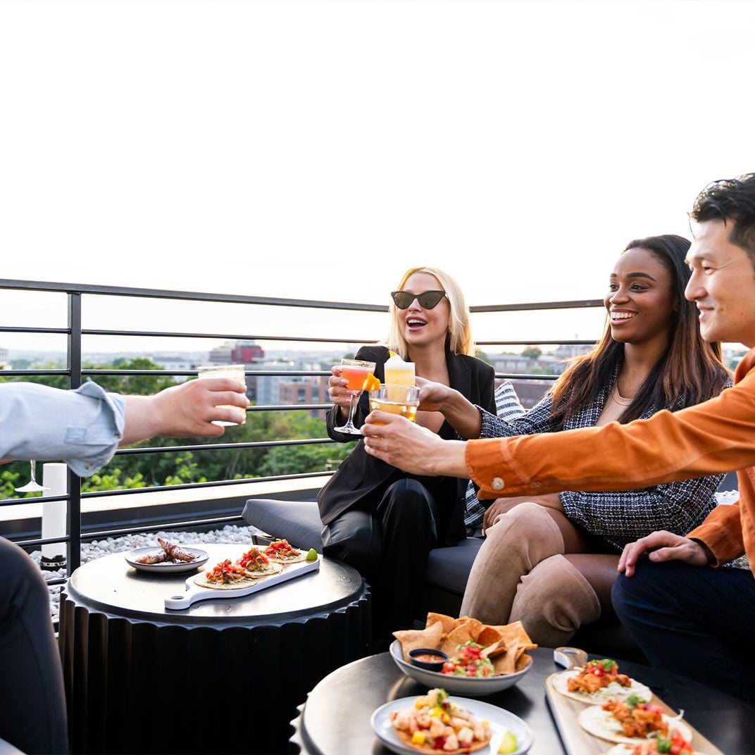Friends toast golden hour cocktails on the rooftop terrace, sharing small plates while the city sprawls beyond the sleek railings in warm evening light.