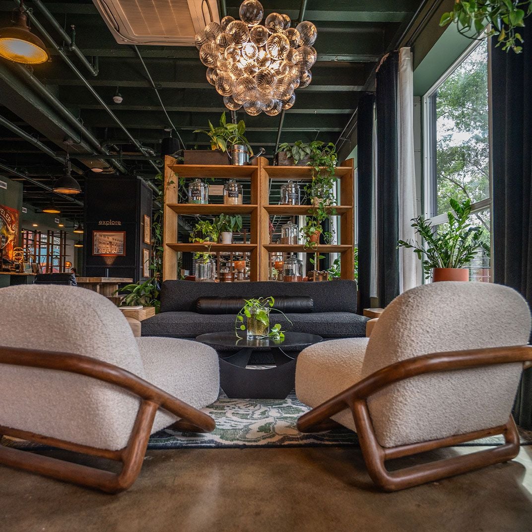 Mid-century armchairs and cascading greenery create an inviting lounge where industrial beams meet warm wood shelving and statement bubble lighting.