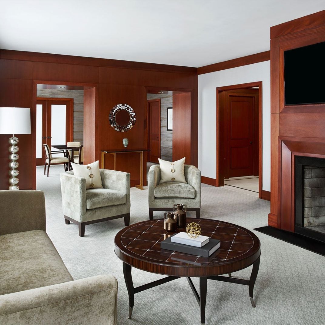 Rich mahogany millwork frames the Presidential Suite's living area, where plush armchairs and a geometric coffee table create an intimate conversation space beside the crackling fireplace.