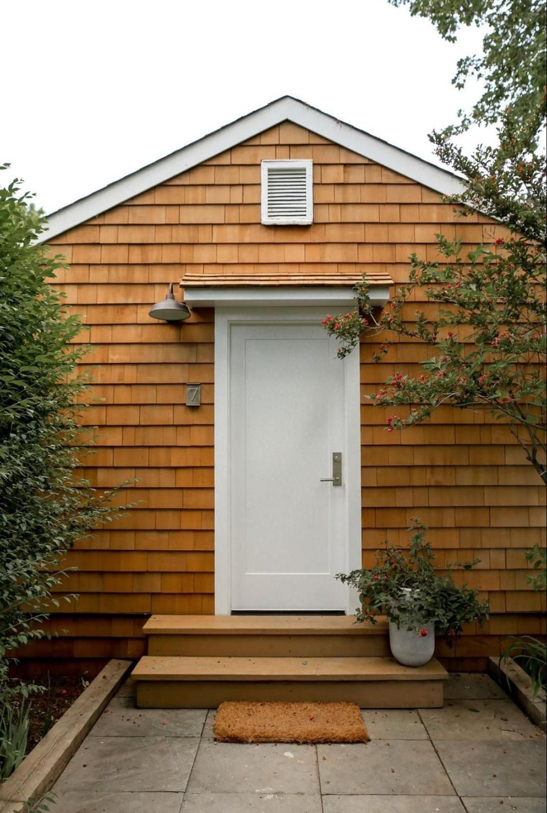 Cedar shingles warm in afternoon light frame a crisp white door, where potted plants and weathered steps create an intimate arrival at this Hamptons retreat.
