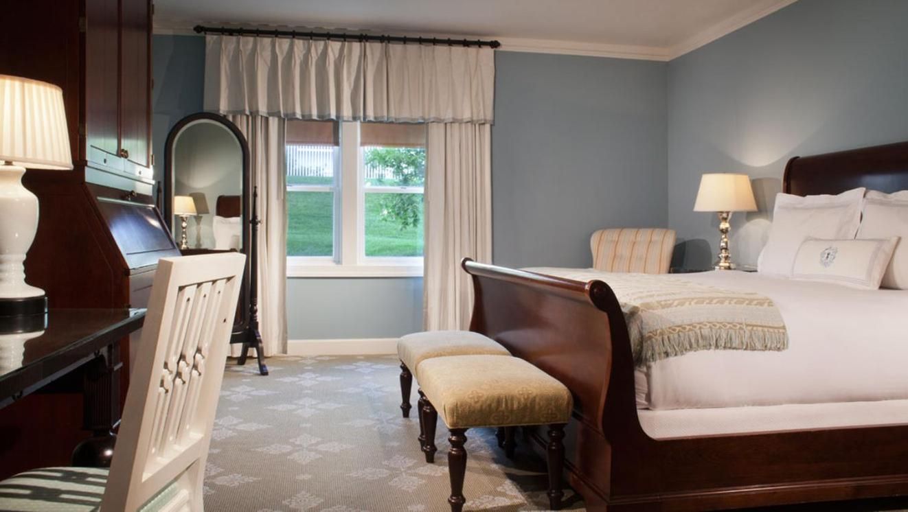 Soft morning light filters through crisp white curtains, illuminating a refined guest room where traditional dark wood furnishings meet soothing blue-gray walls and plush neutral linens.