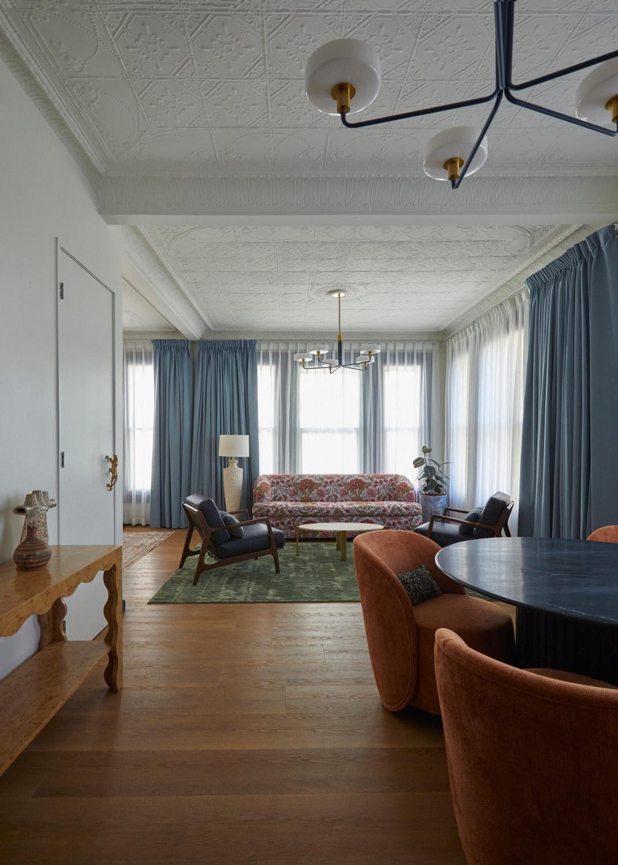 Ornate tin ceilings and jewel-toned curtains frame this sophisticated living space, where mid-century furniture mingles with vintage charm beneath dramatic modern lighting.
