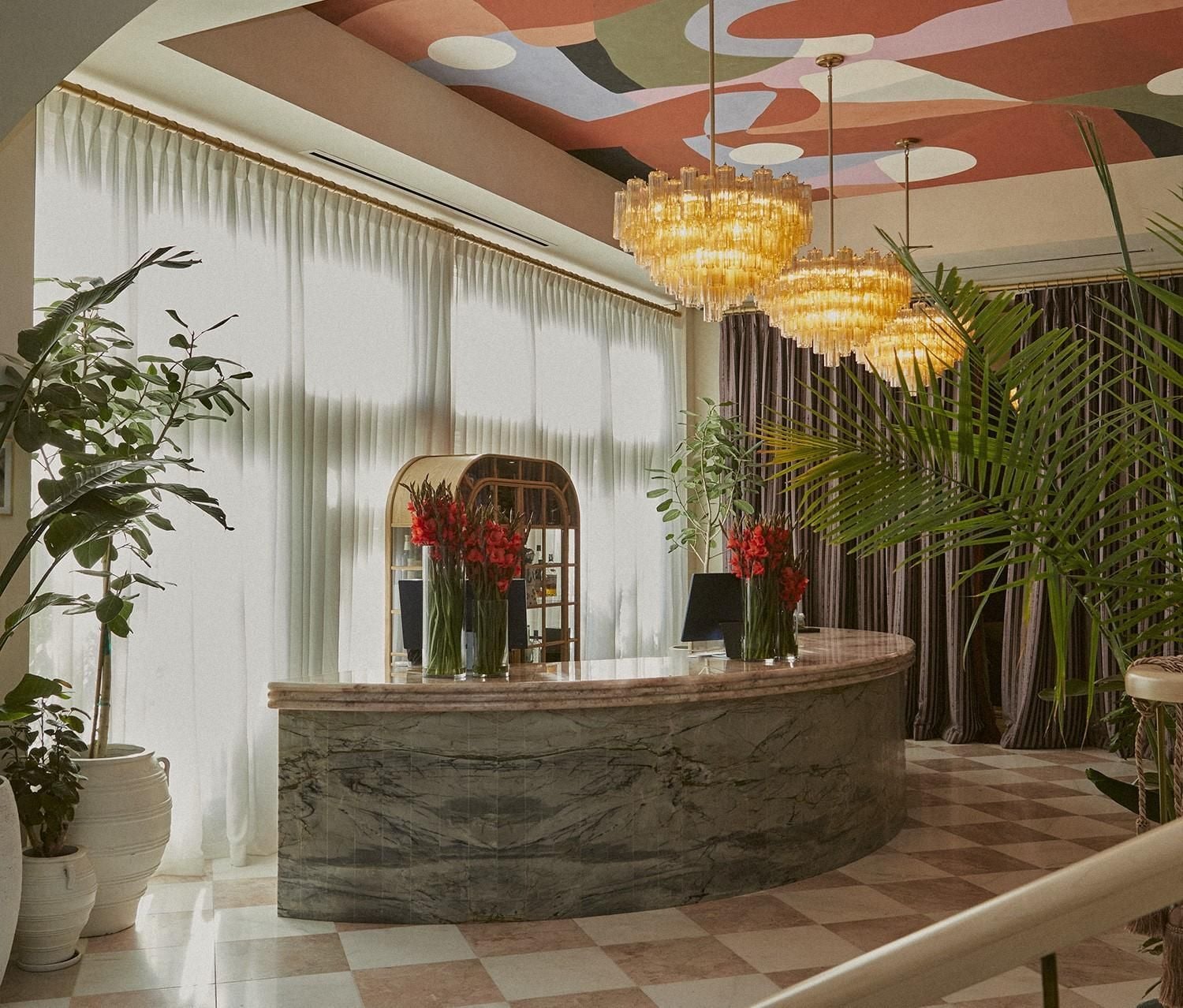 Curved marble reception desk anchors the vibrant lobby, where golden chandeliers cast warm light across painted ceiling murals and lush tropical greenery.