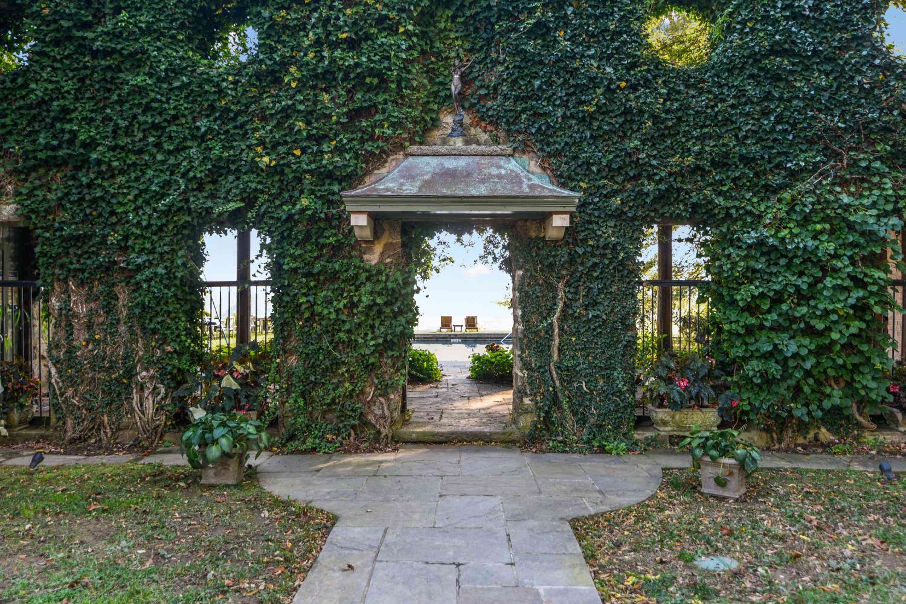 Ivy-draped stone arches frame a romantic passage through manicured gardens, where weathered flagstones lead toward sunlit terraces and distant mountain views.