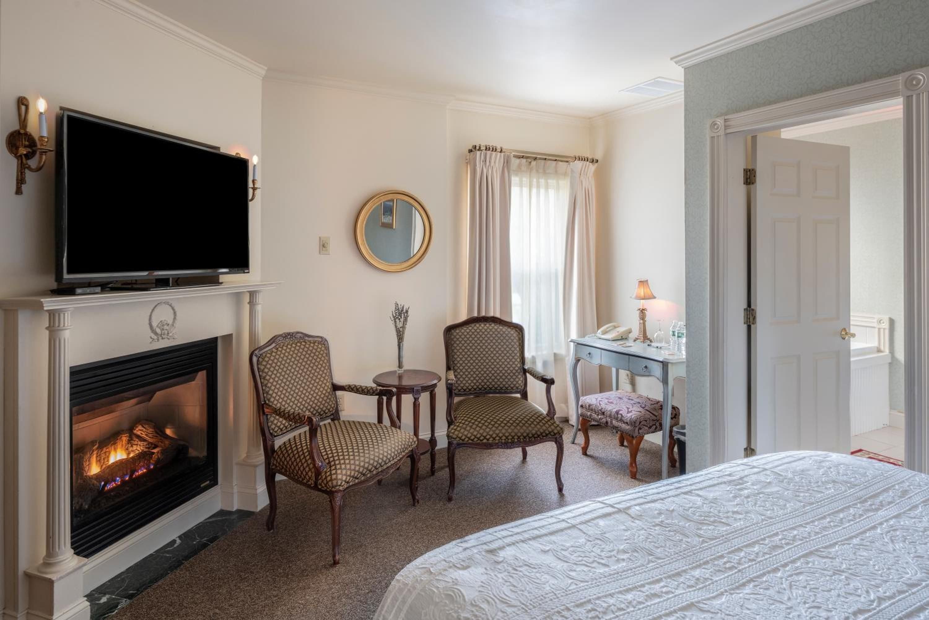 Crackling flames cast a warm glow across patterned armchairs and plush bedding, where colonial elegance meets intimate comfort in this fireside sanctuary.