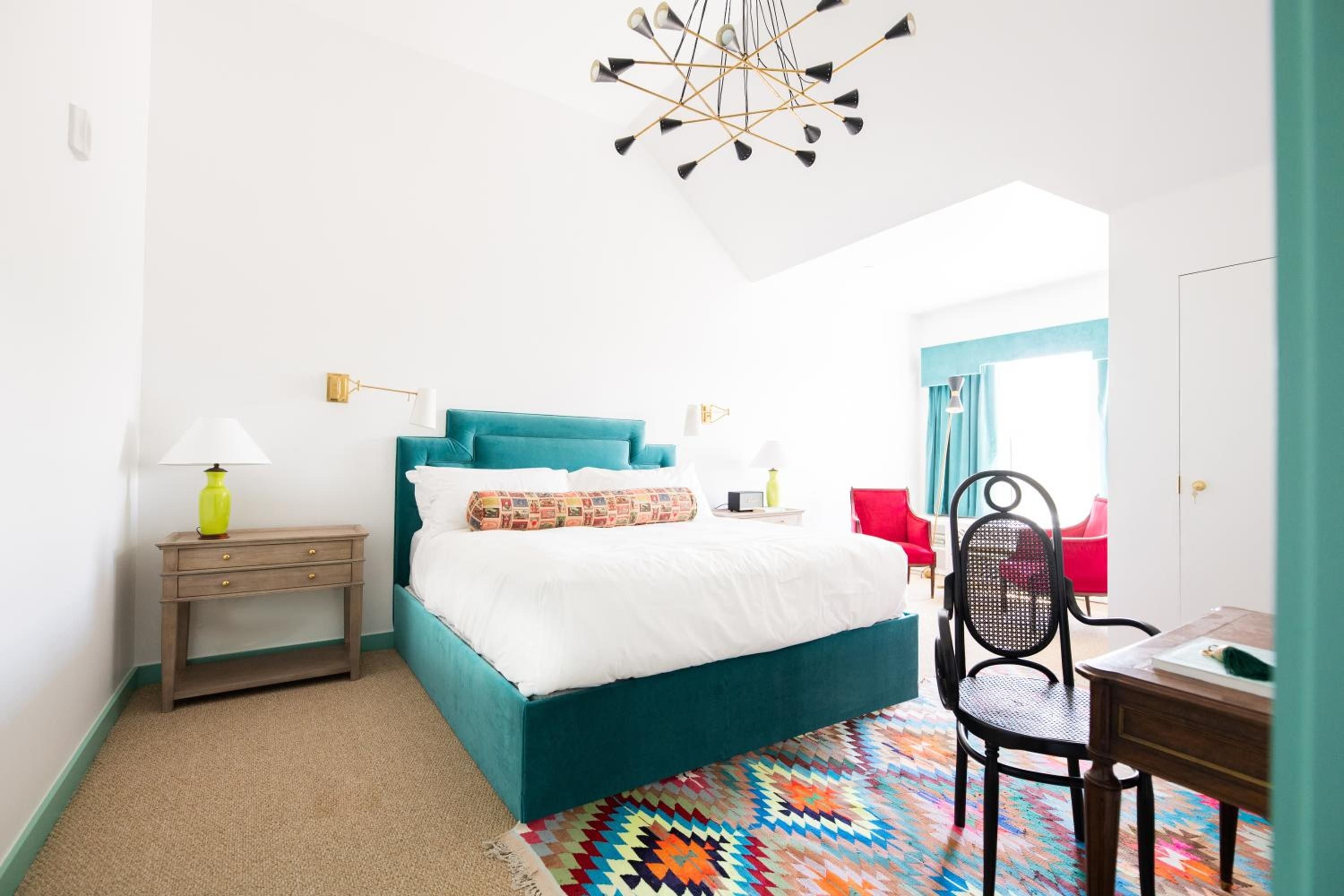 Teal upholstered beds anchor bright guest rooms where geometric kilim rugs pop against neutral sisal, while brass swing-arm sconces cast warm pooled light.