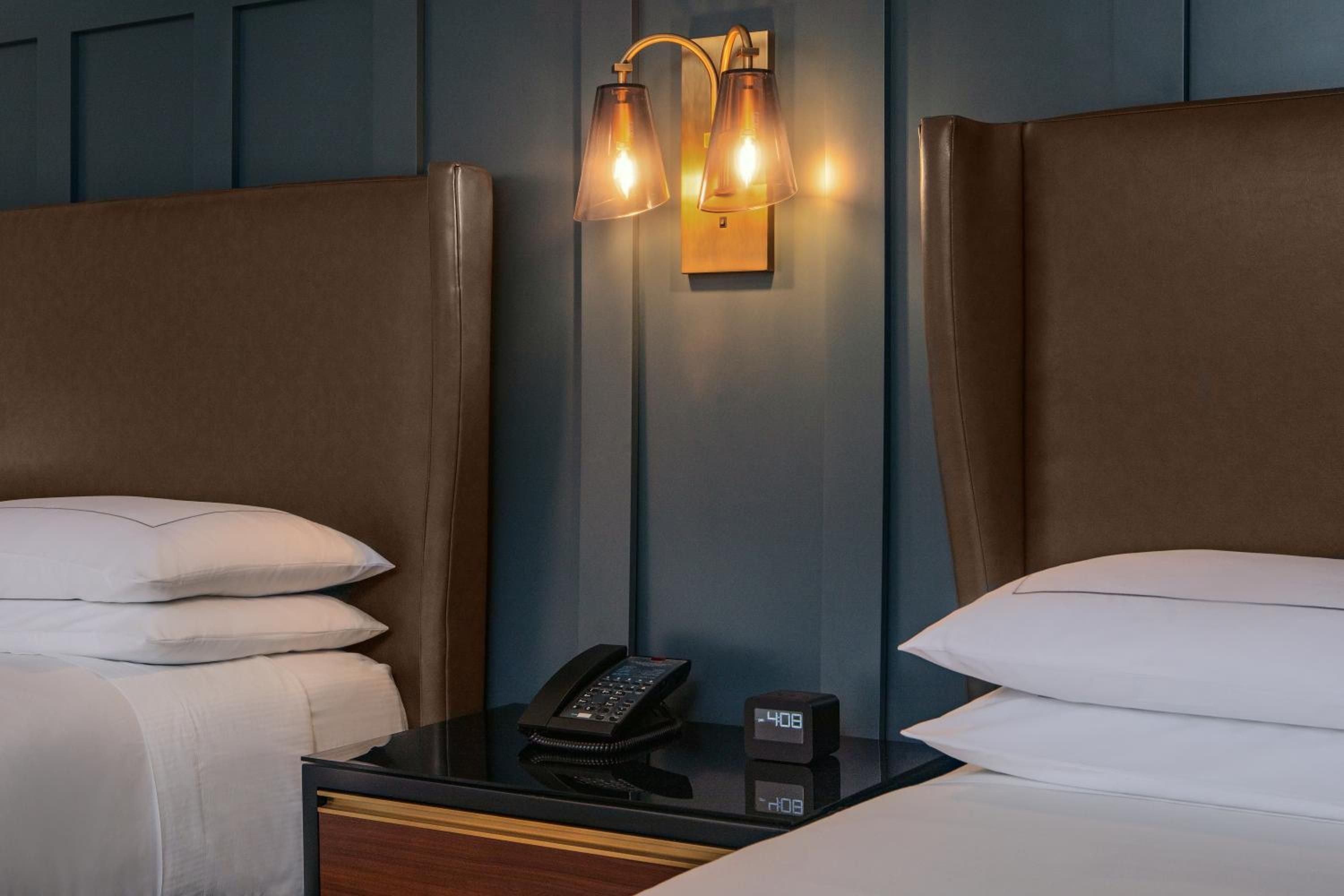 Warm brass sconces cast amber light between twin beds, while a sleek nightstand holds modern amenities in this thoughtfully appointed guest room.