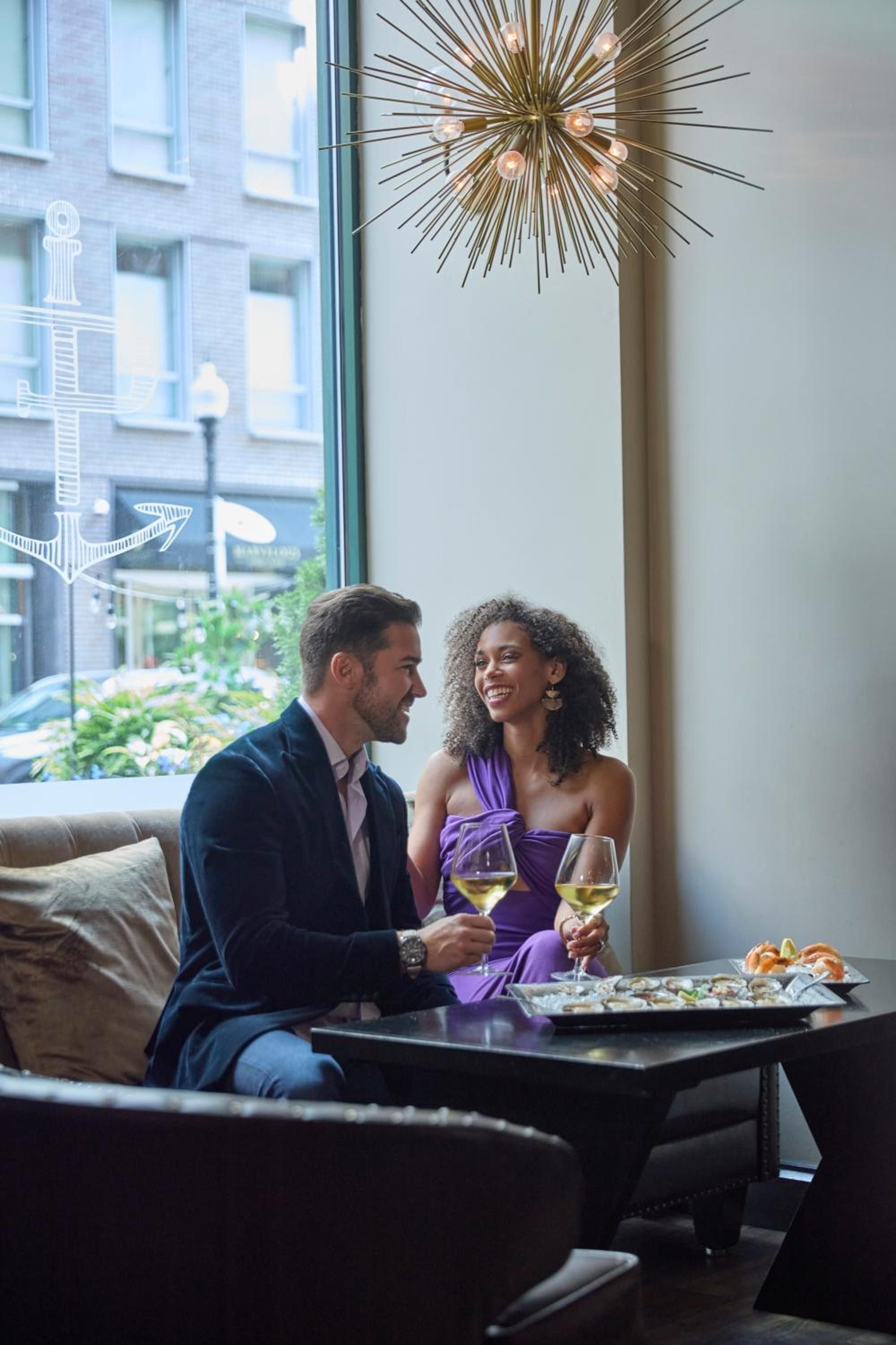 Intimate dining unfolds beneath a striking starburst chandelier, where couples savor wine and conversation against floor-to-ceiling windows framing Boston's vibrant streetscape.