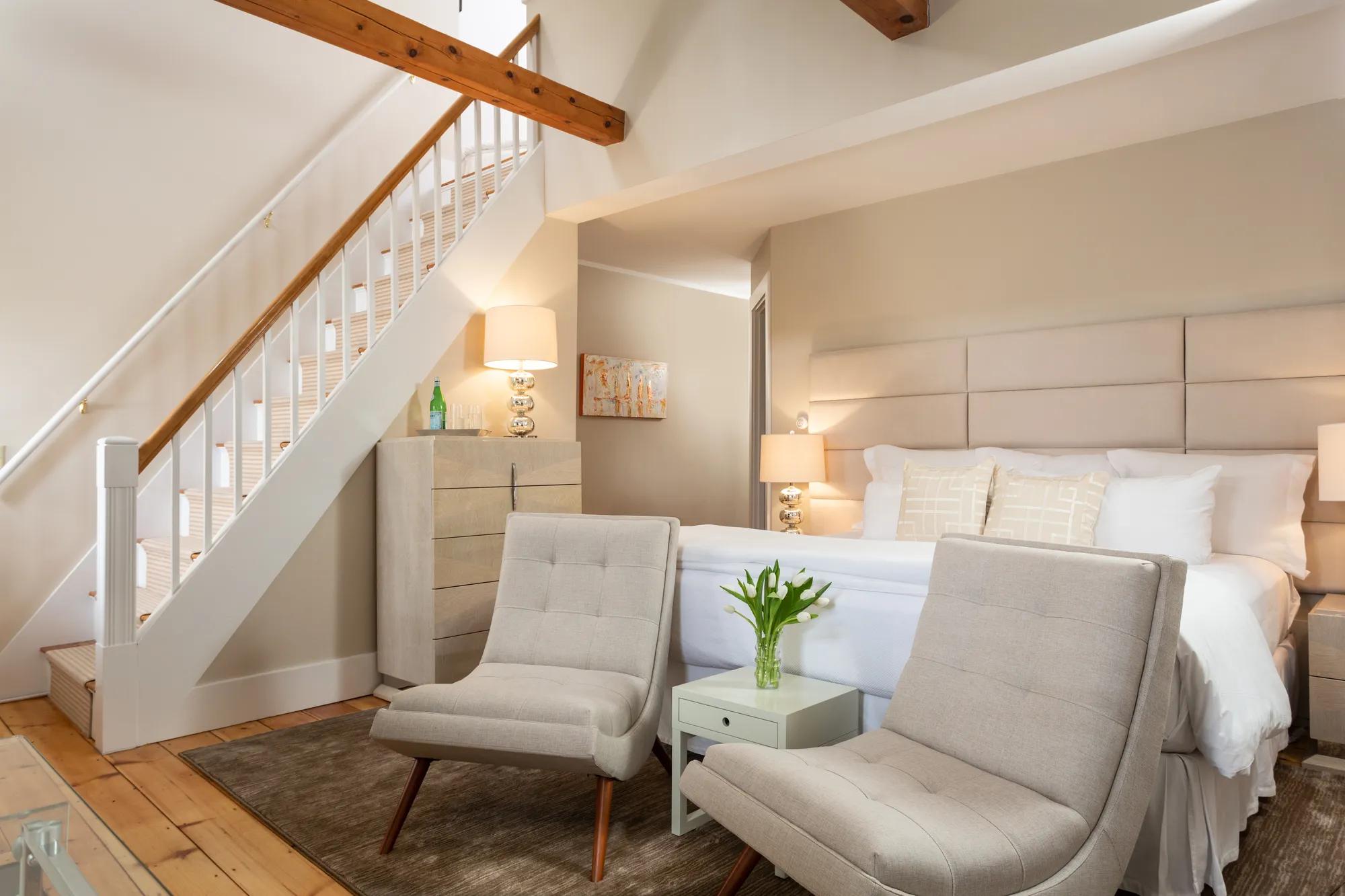 Exposed beams and whitewashed walls frame a cozy seating area beneath the staircase, where morning light filters through creating intimate shadows and warm textures.