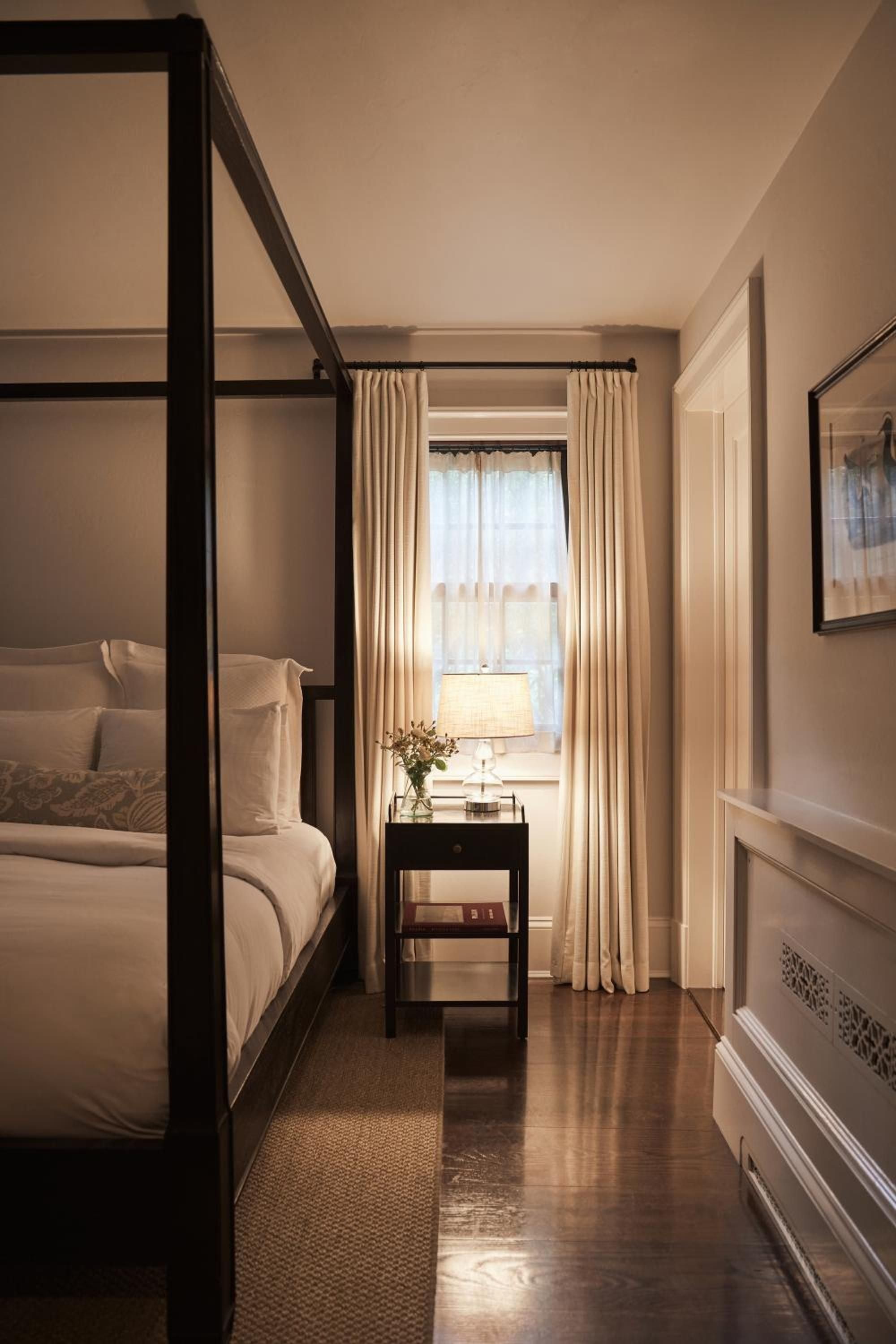 Candlelight flickers across polished hardwood floors in this intimate guest room, where dark four-poster beds meet cream linens and golden curtains frame peaceful garden views.