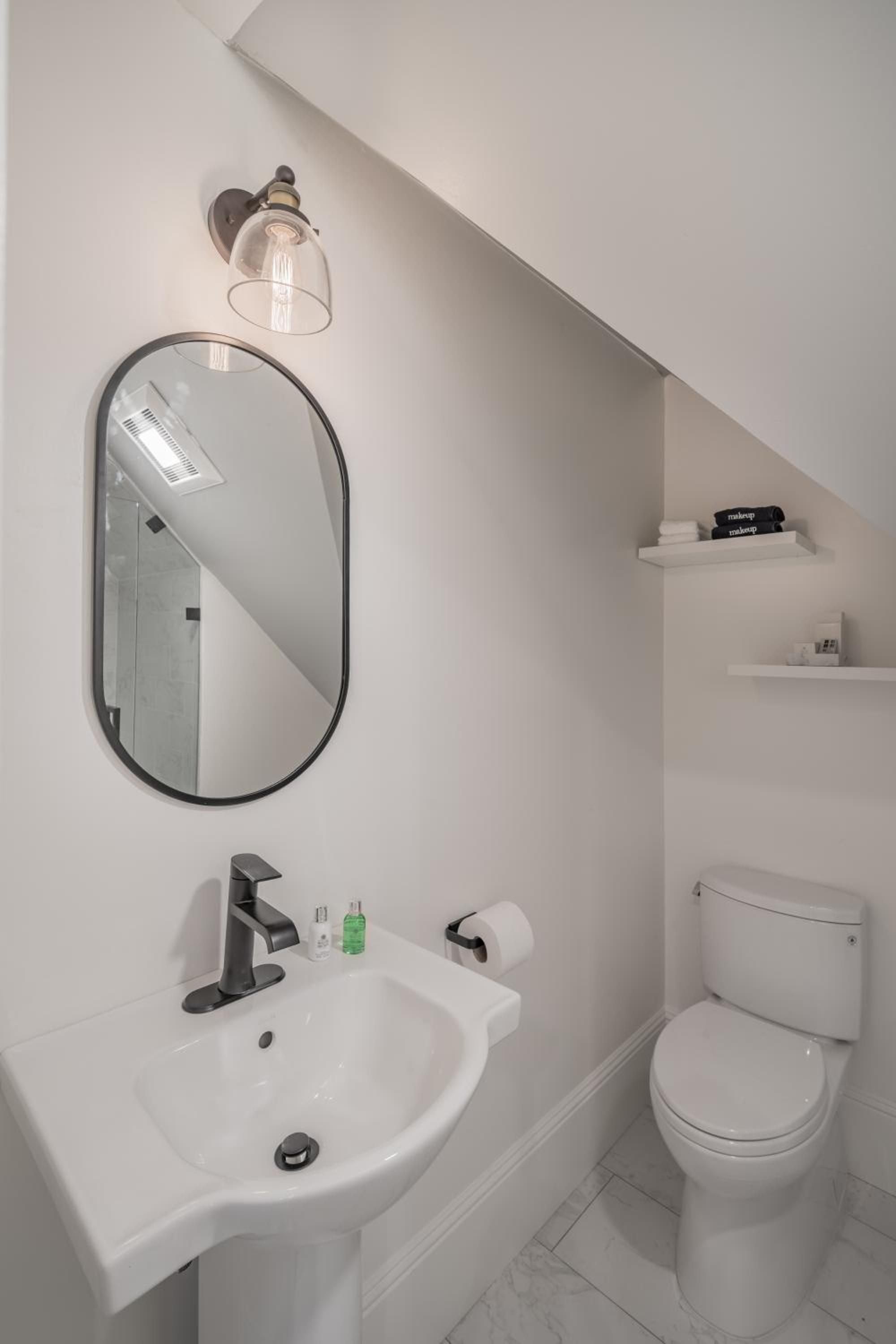 Clever design maximizes this compact powder room with an oval mirror, crisp white pedestal sink, and floating shelves tucked beneath the slanted ceiling.