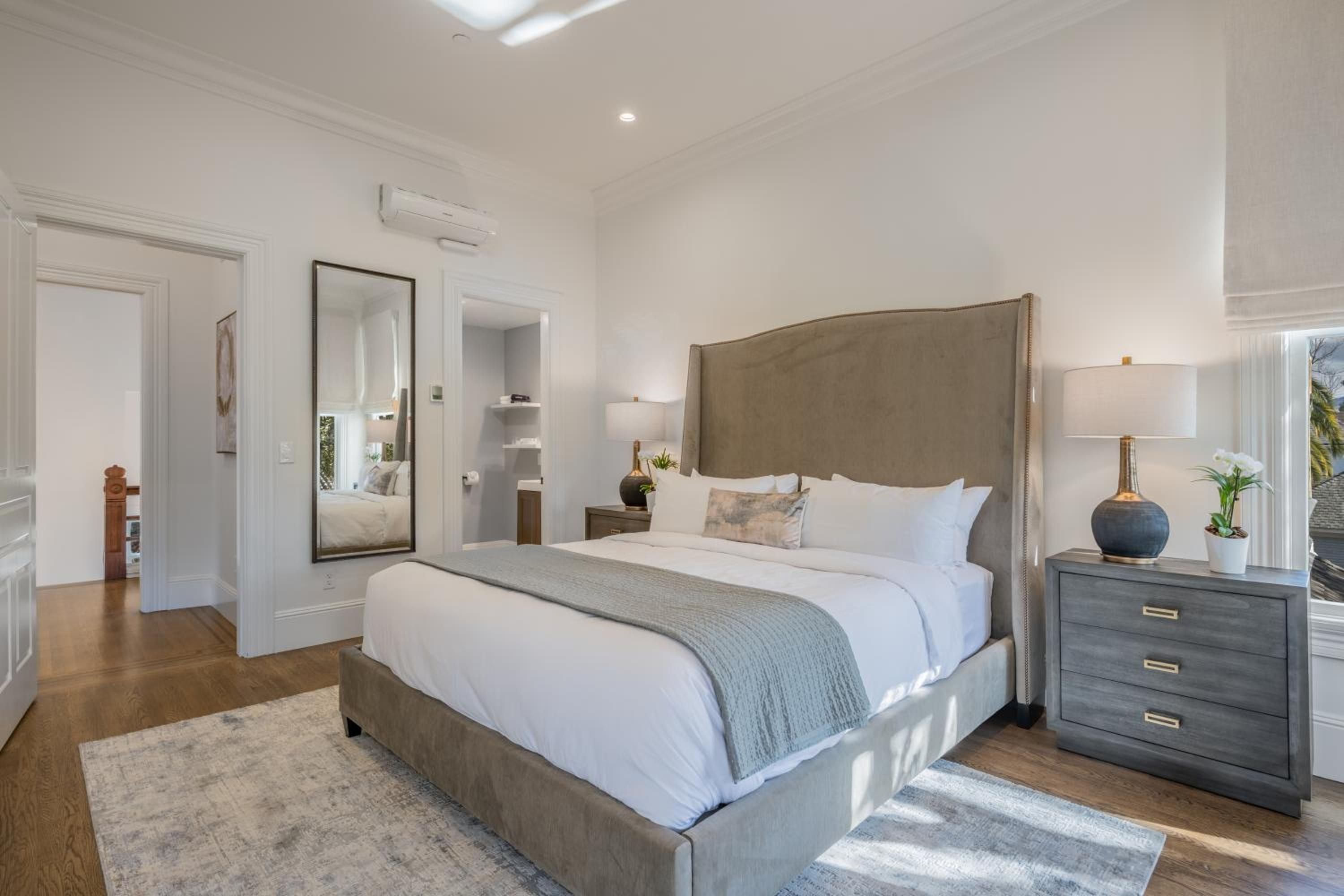 Soft morning light filters across polished hardwood floors, illuminating crisp white linens and the room's sophisticated taupe headboard in this serene guest suite.