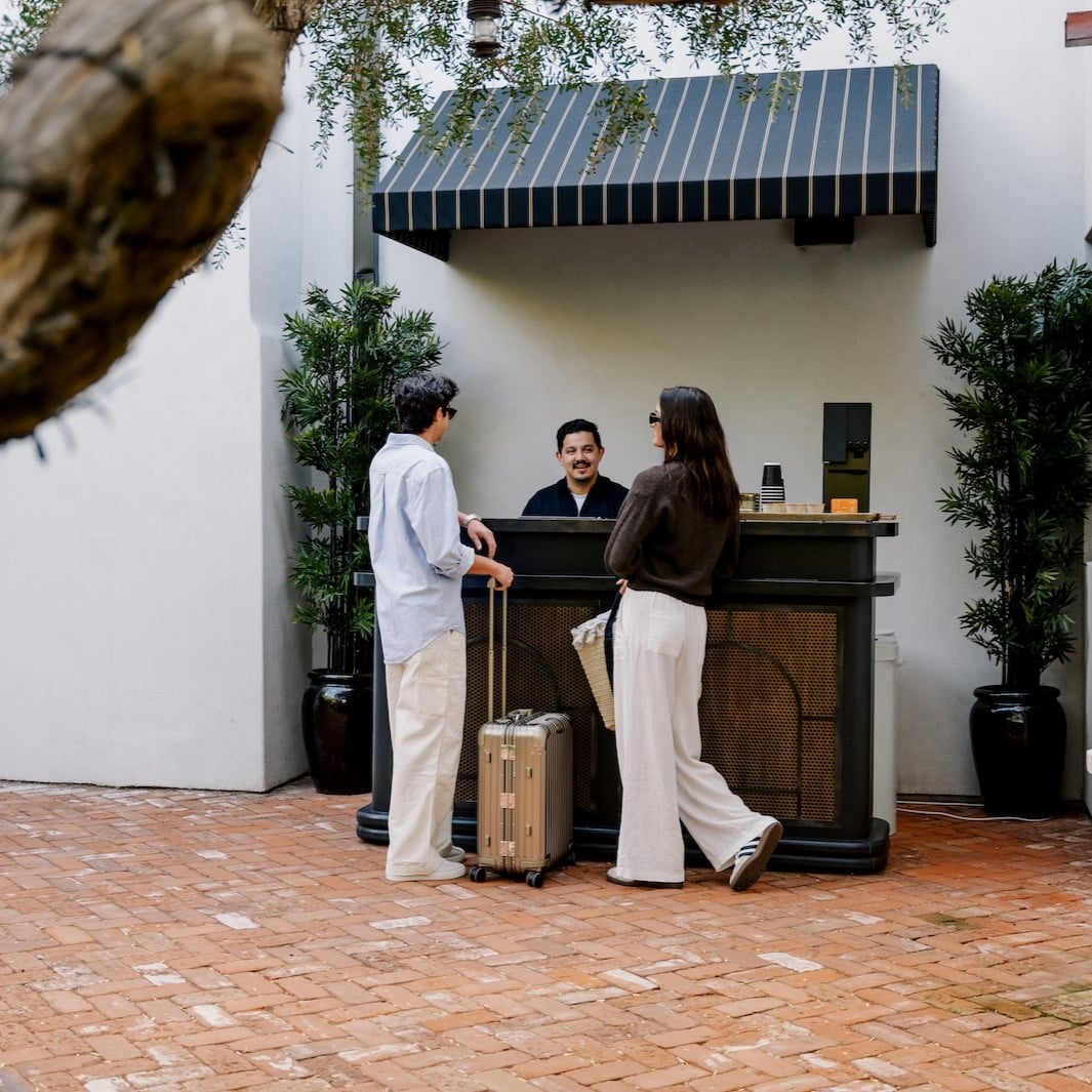 Guests gather at the outdoor coffee cart beneath a striped awning, where artisanal brews meet California's casual elegance on sun-warmed brick pathways.