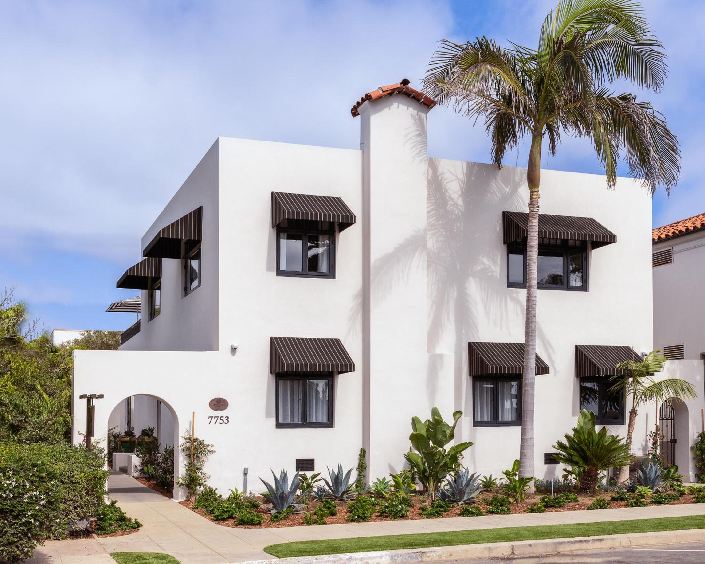 Clean white stucco walls and terra cotta accents define this Spanish Colonial Revival residence, where striped awnings frame windows beneath towering palms and azure skies.