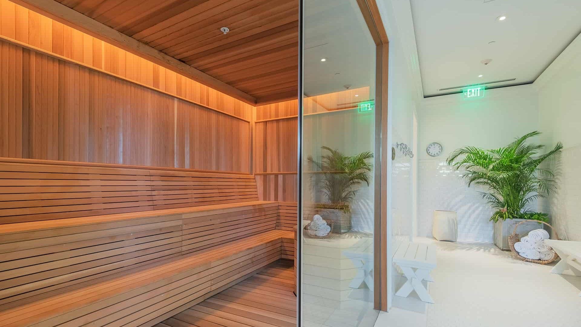 Cedar-lined sauna chambers glow with amber warmth beside marble relaxation areas, where plush towels and tropical palms create an intimate wellness sanctuary.