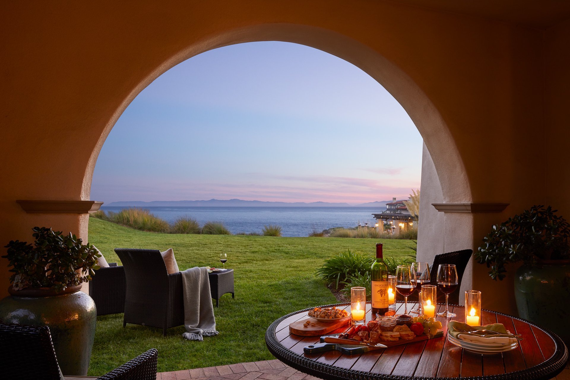 Candlelit dining beneath Mediterranean arches frames endless ocean vistas, where wine glasses catch the golden hour light and distant islands float on the horizon.