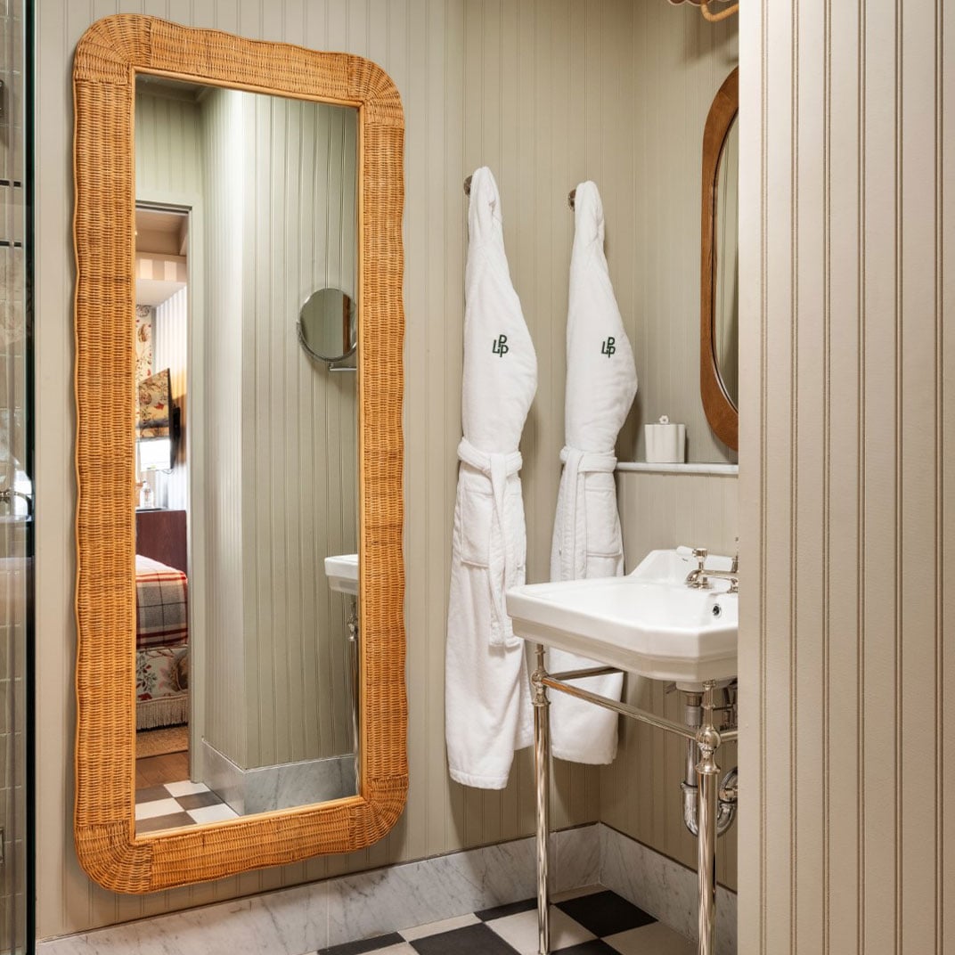 Wicker-framed mirrors and plush monogrammed robes create a cozy, residential feel in thoughtfully appointed bathrooms with checkered marble floors and vintage-inspired fixtures.