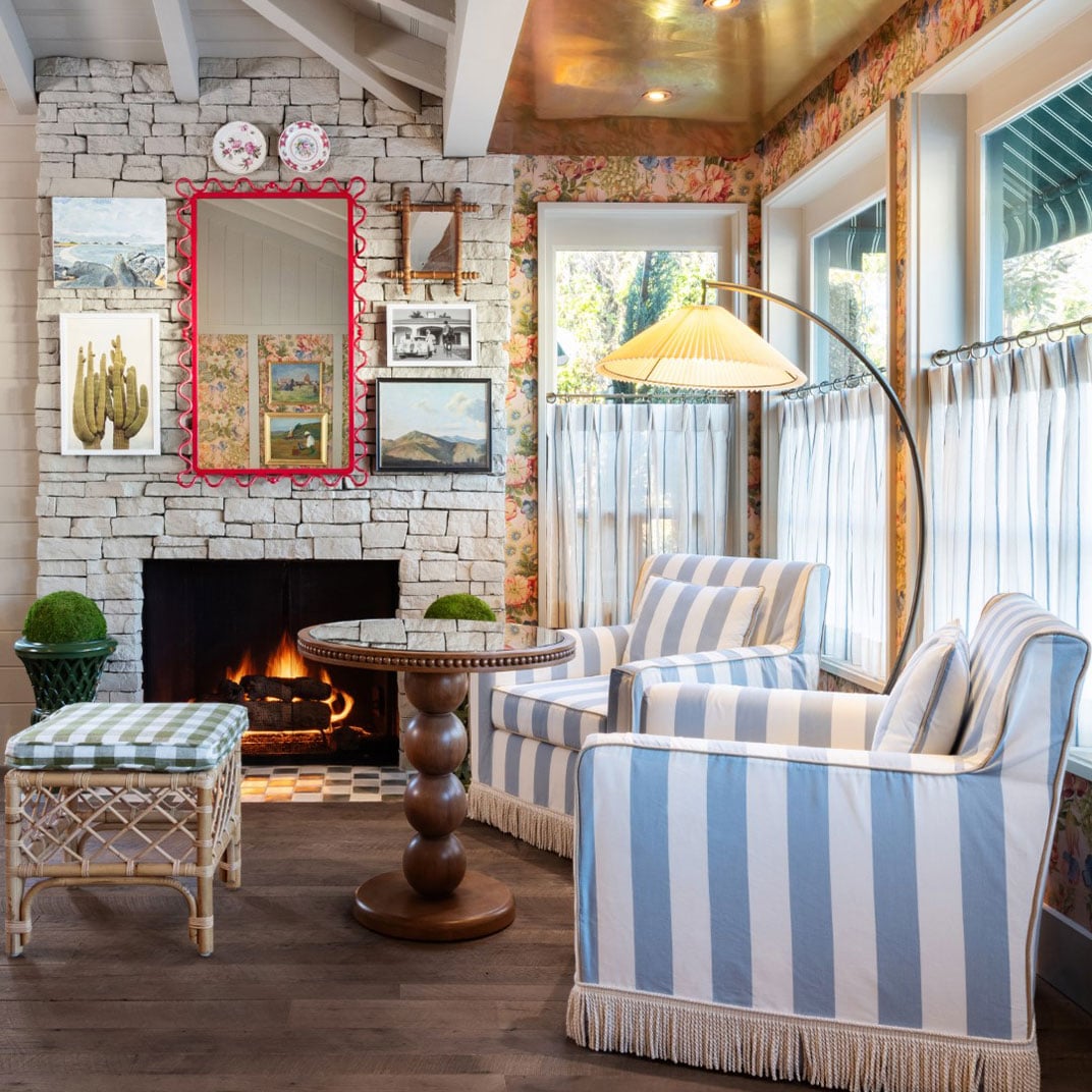 Blue-striped armchairs gather around a crackling stone fireplace, while vintage landscape paintings and floral wallpaper create an intimate, cottage-like retreat.