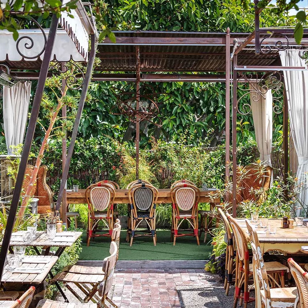 Wicker bistro chairs cluster beneath iron pergolas draped in flowing curtains, creating intimate dining nooks amid lush greenery and dappled sunlight.