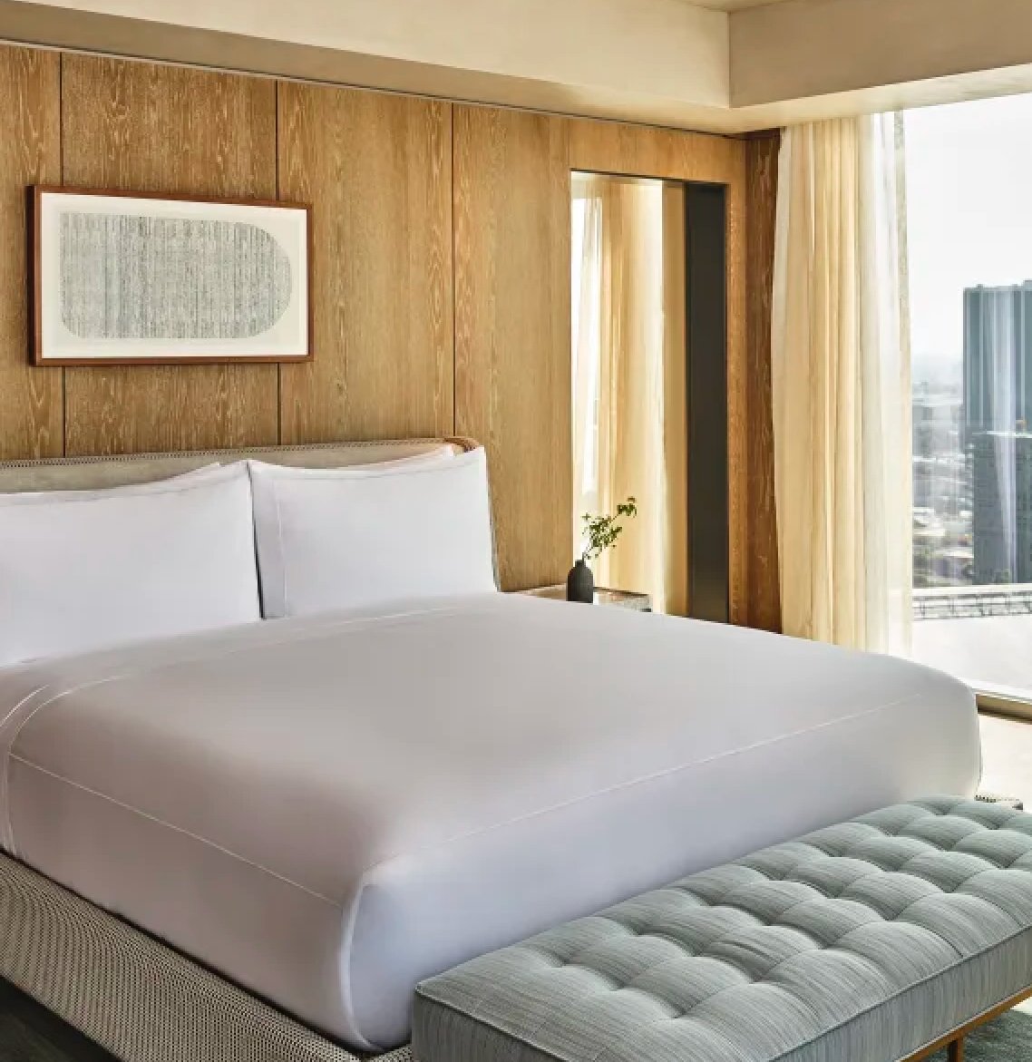 Warm oak paneling and crisp white linens create a serene sanctuary, while floor-to-ceiling windows frame downtown LA's gleaming skyline beyond flowing curtains.