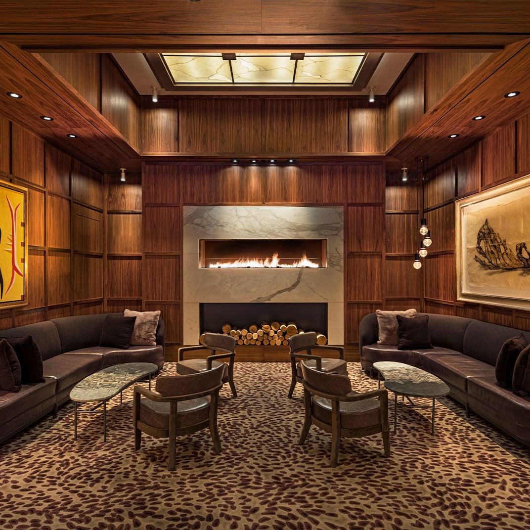 Rich walnut paneling and marble fireplace create an intimate sanctuary where guests sink into plush seating beneath a glowing coffered ceiling.