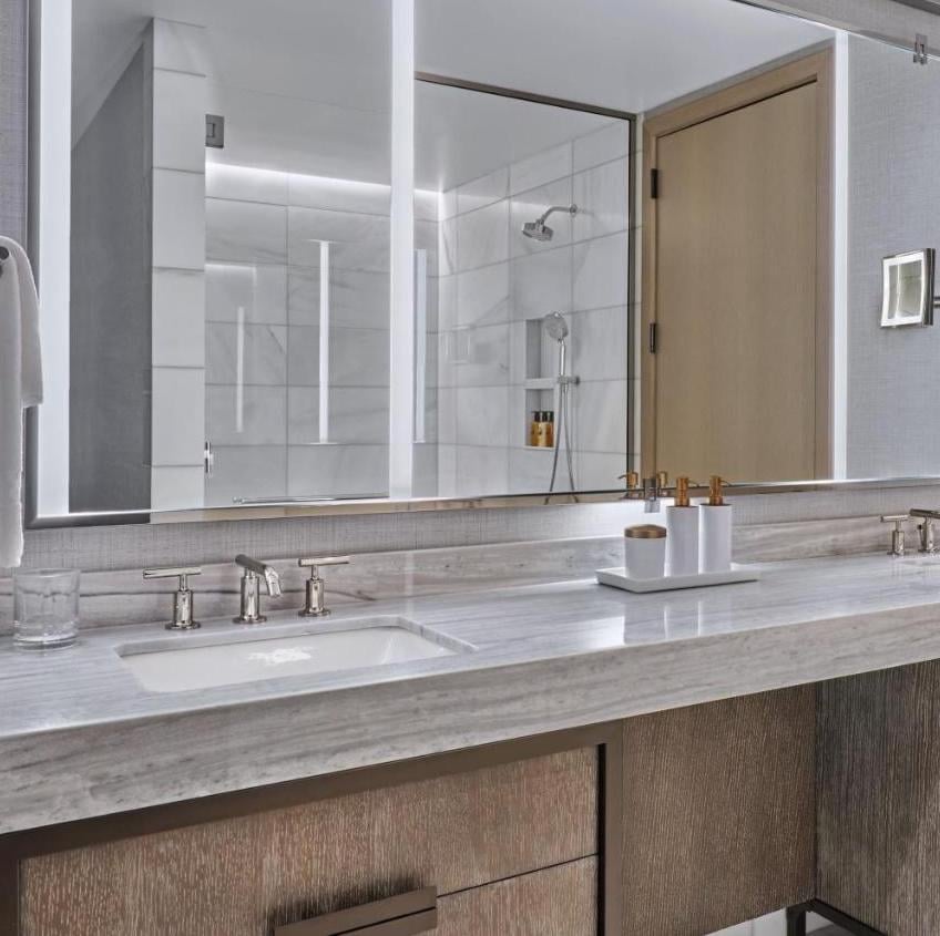 Polished marble counters and warm wood cabinetry create a spa-like retreat, while oversized mirrors reflect the glass-enclosed rainfall shower beyond.