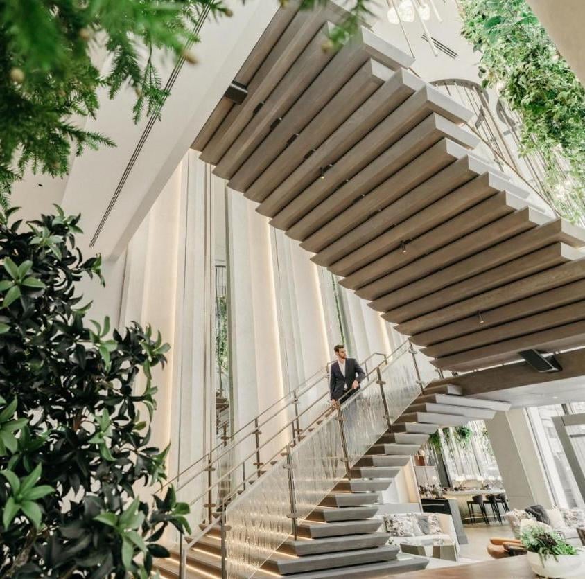 Floating wooden steps ascend beneath a sculptural slatted ceiling, where natural light filters through floor-to-ceiling windows and lush greenery softens modern lines.