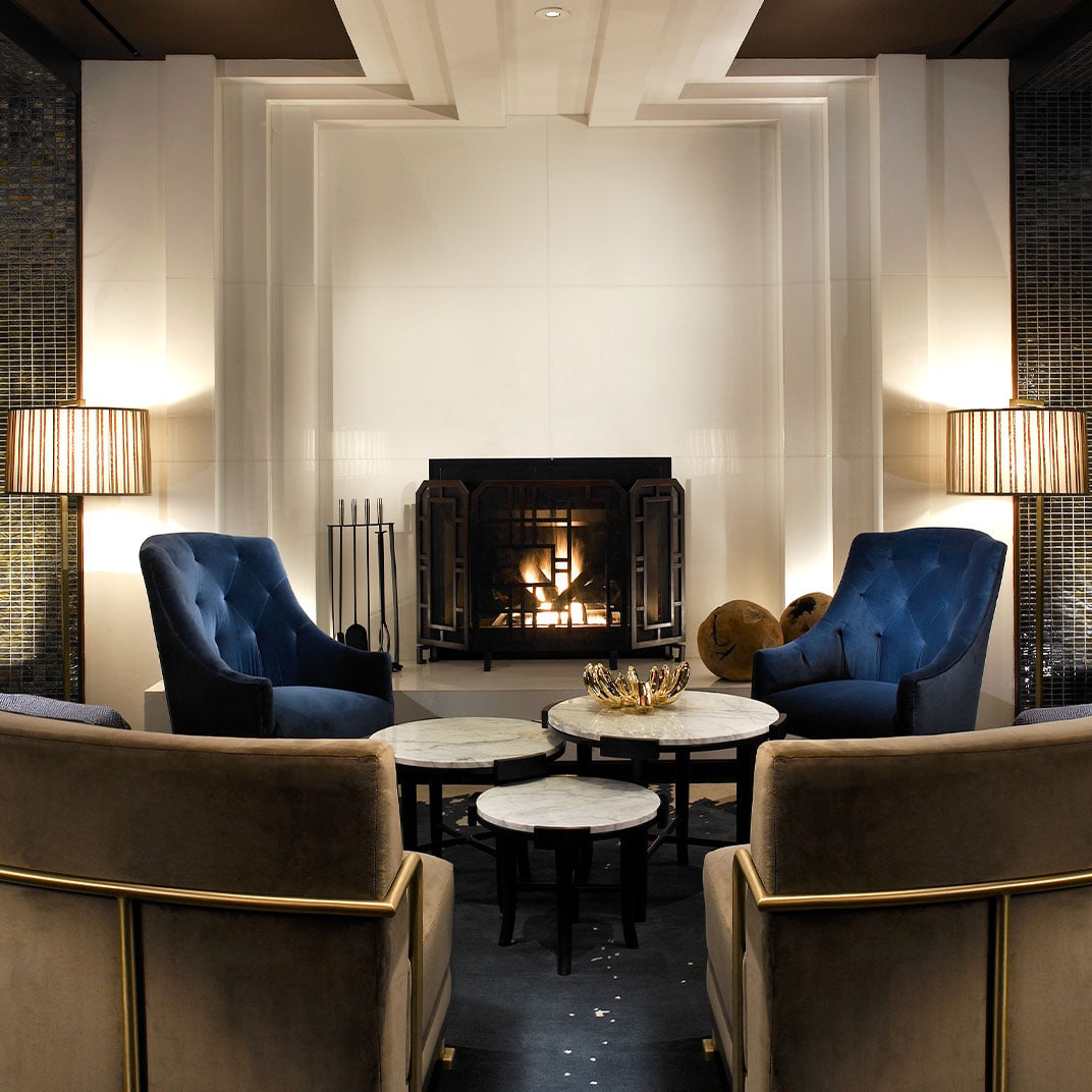 Velvet navy armchairs and gleaming marble tables create an intimate conversation circle around the crackling fireplace, where warm amber light dances across coffered ceilings.