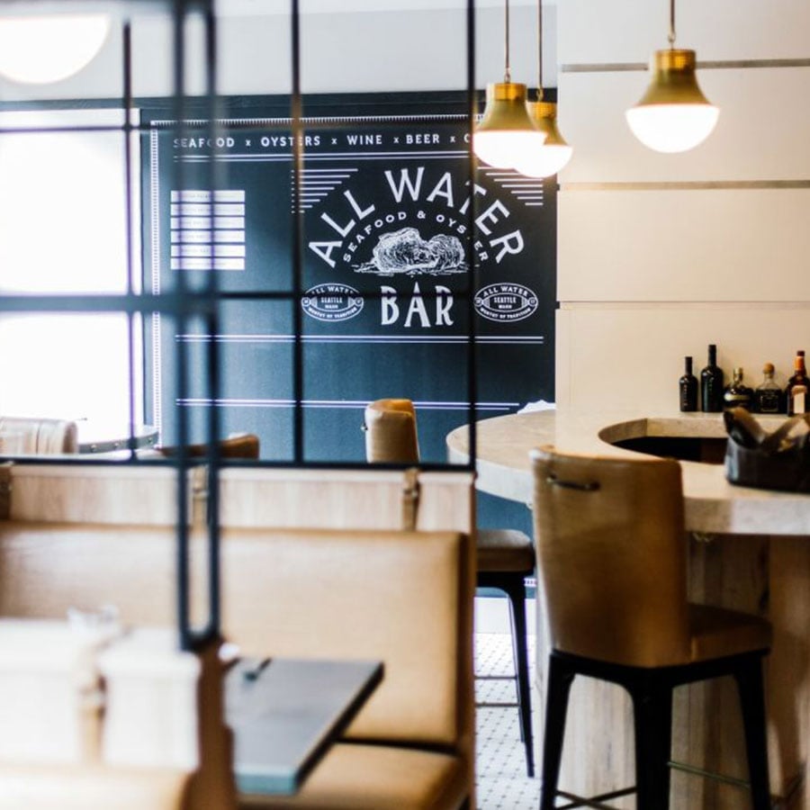 Golden pendant lights cast a warm glow over leather bar stools at All Water Seafood & Oyster Bar, where maritime-themed signage hints at fresh Pacific Northwest catches.