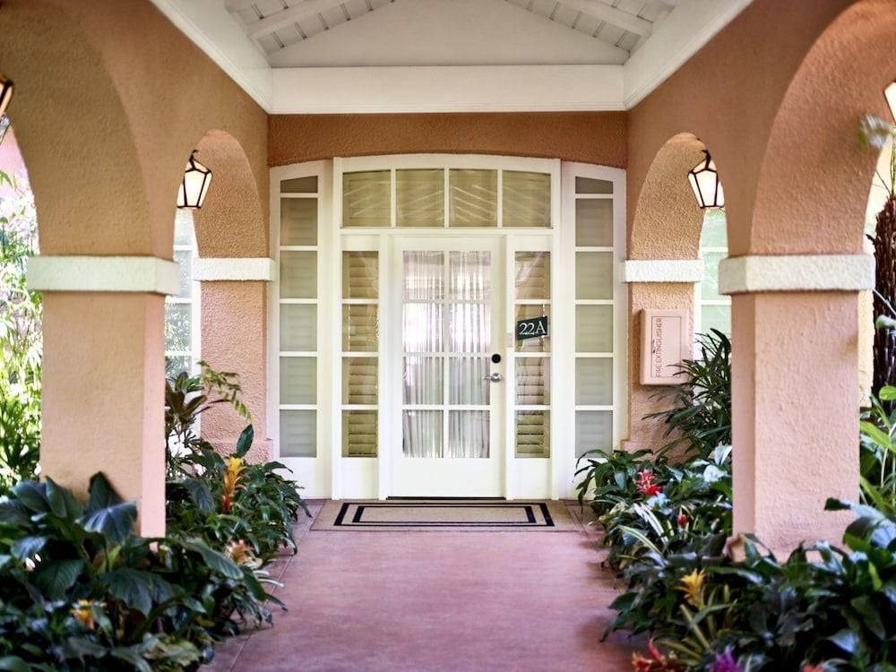 Lush tropical plantings frame the welcoming entrance to suite 22A, where coral-hued stucco walls and white columns create an intimate residential feeling beneath soaring arched ceilings.