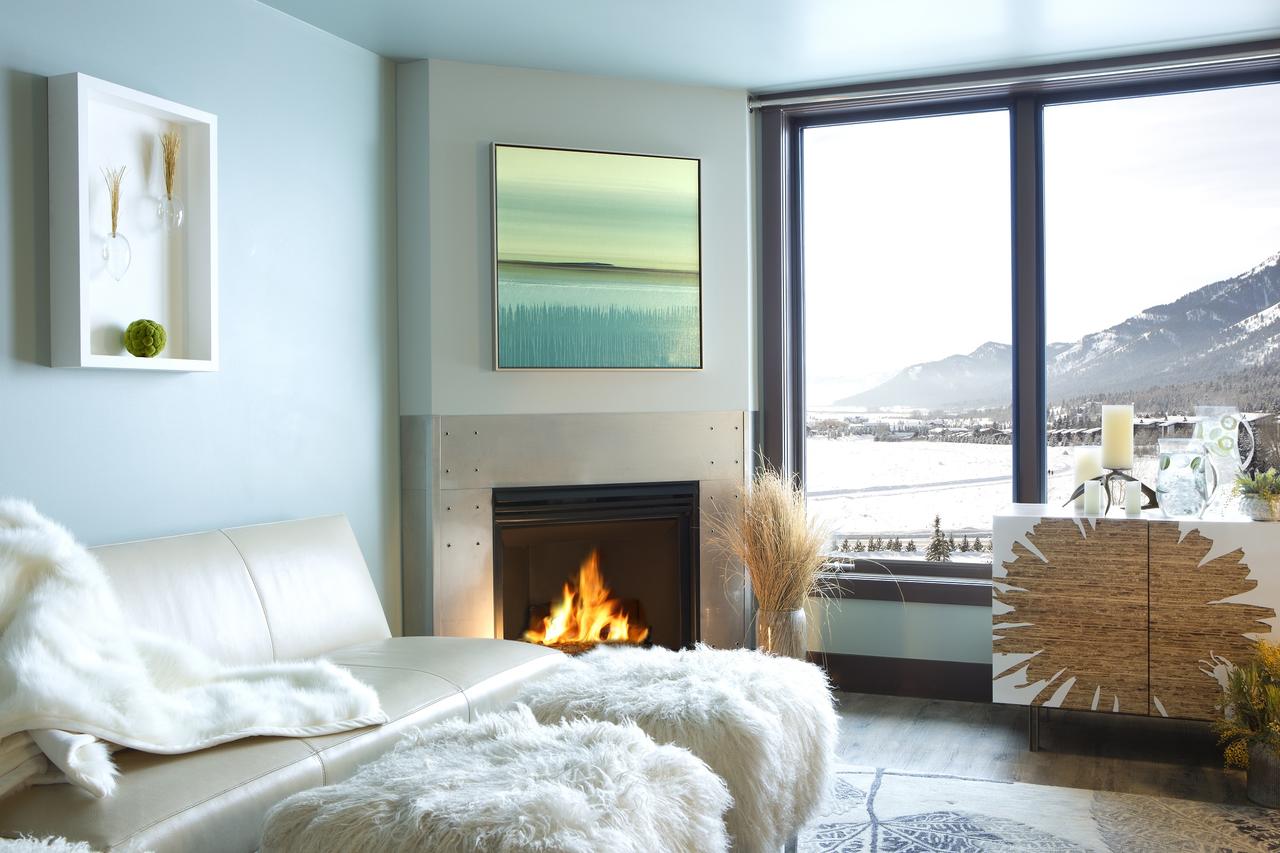 White leather seating and fluffy throws invite lingering by the crackling fireplace while snow-dusted mountains frame this serene alpine retreat through floor-to-ceiling windows.