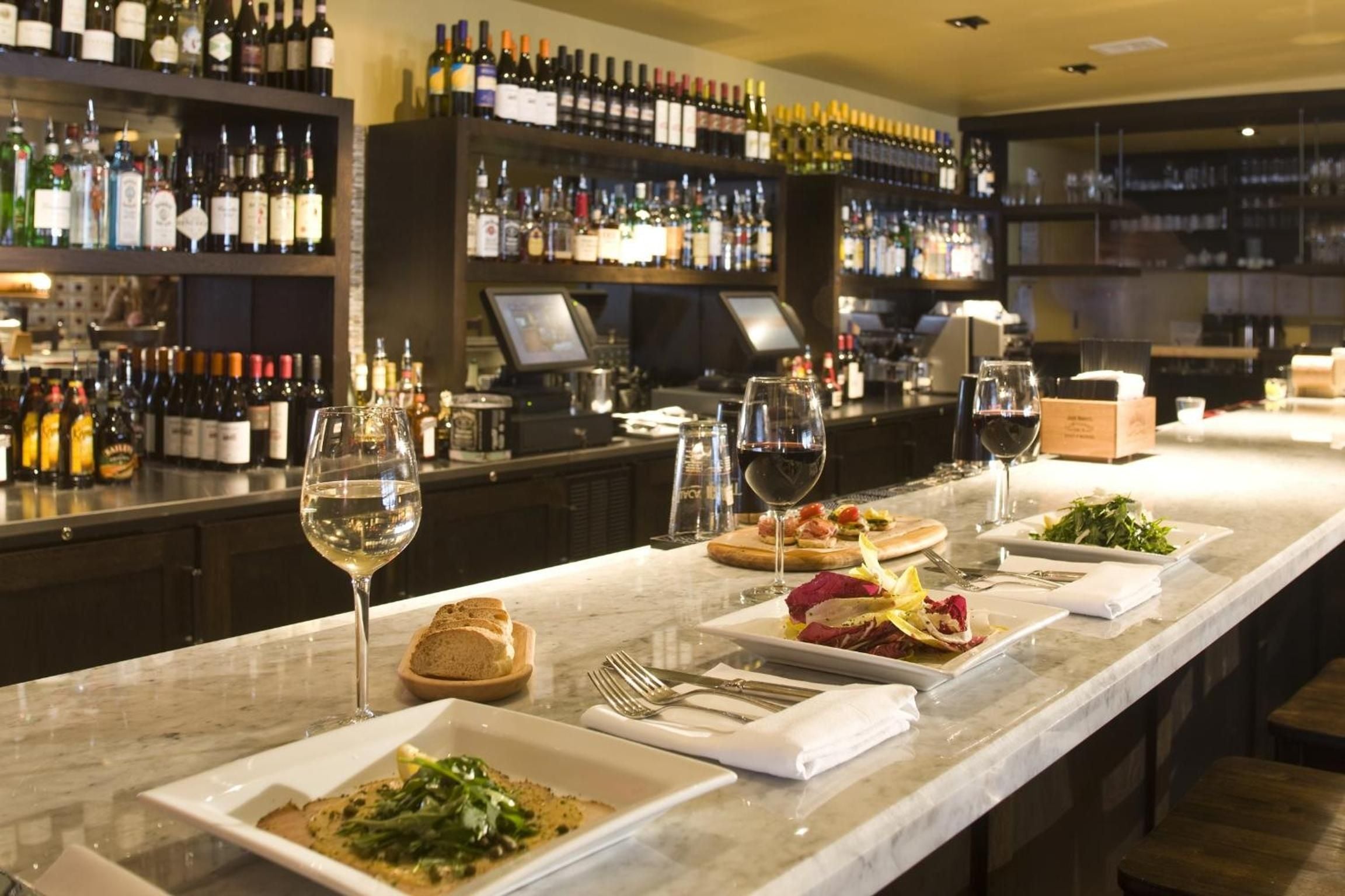 Marble countertops gleam beneath towering wine displays at the sophisticated bar, where artisanal plates and perfectly paired glasses create an intimate tasting experience.