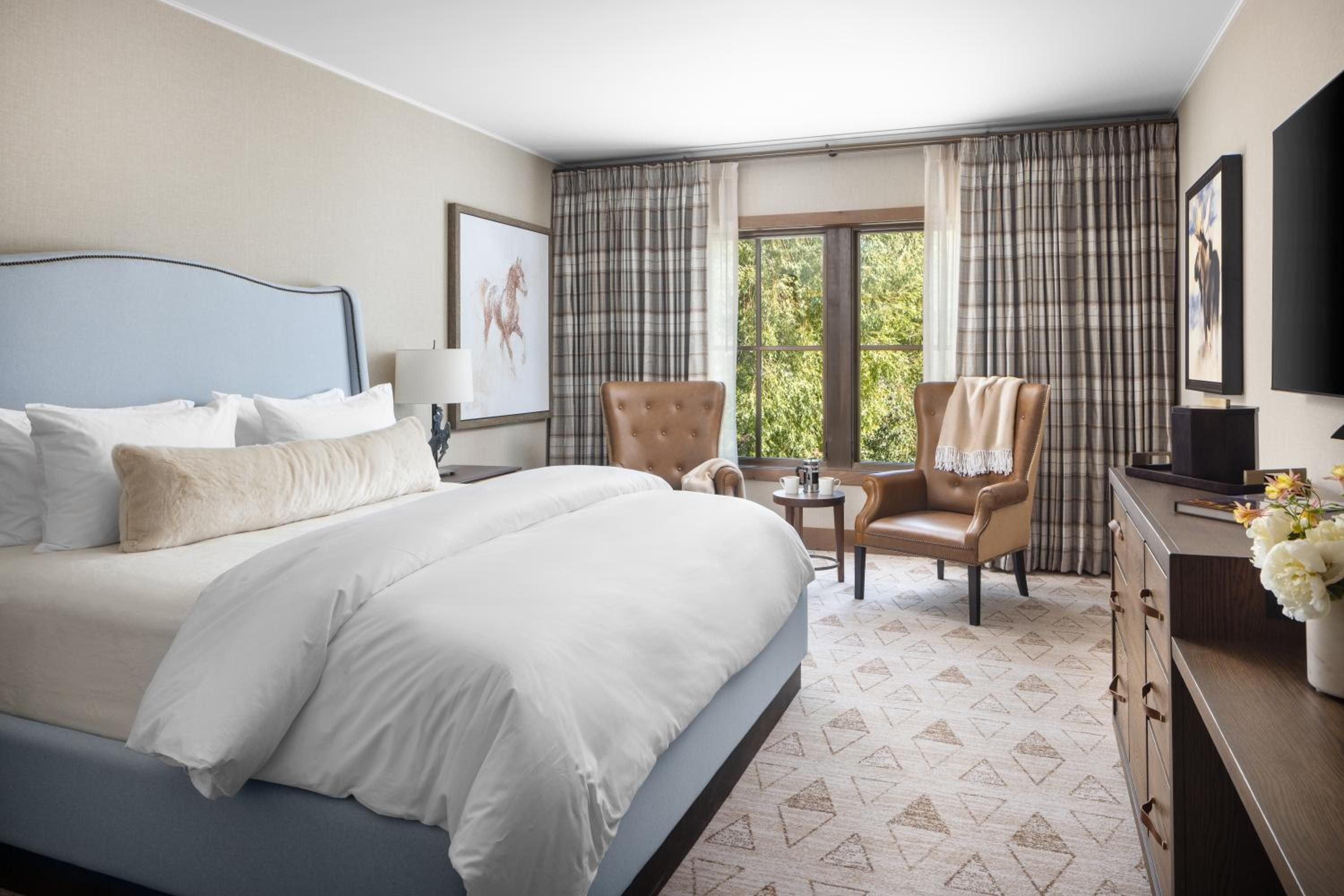 Soft blue linens and leather wingback chairs create an intimate retreat, while plaid curtains frame mountain views beyond the generous windows.