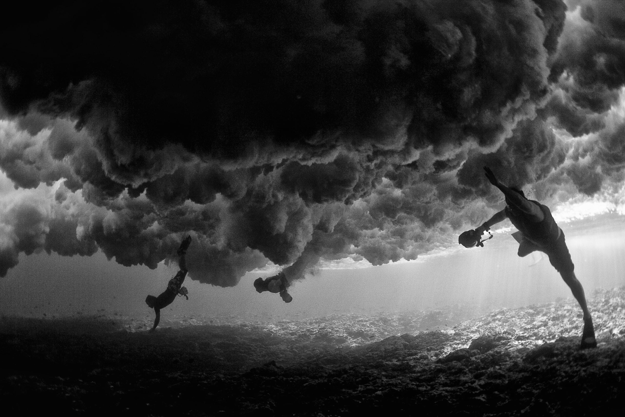 Beneath crystalline waters, silhouettes of swimmers create an ethereal underwater ballet, their forms suspended like graceful shadows against churning bubbles and filtered light.