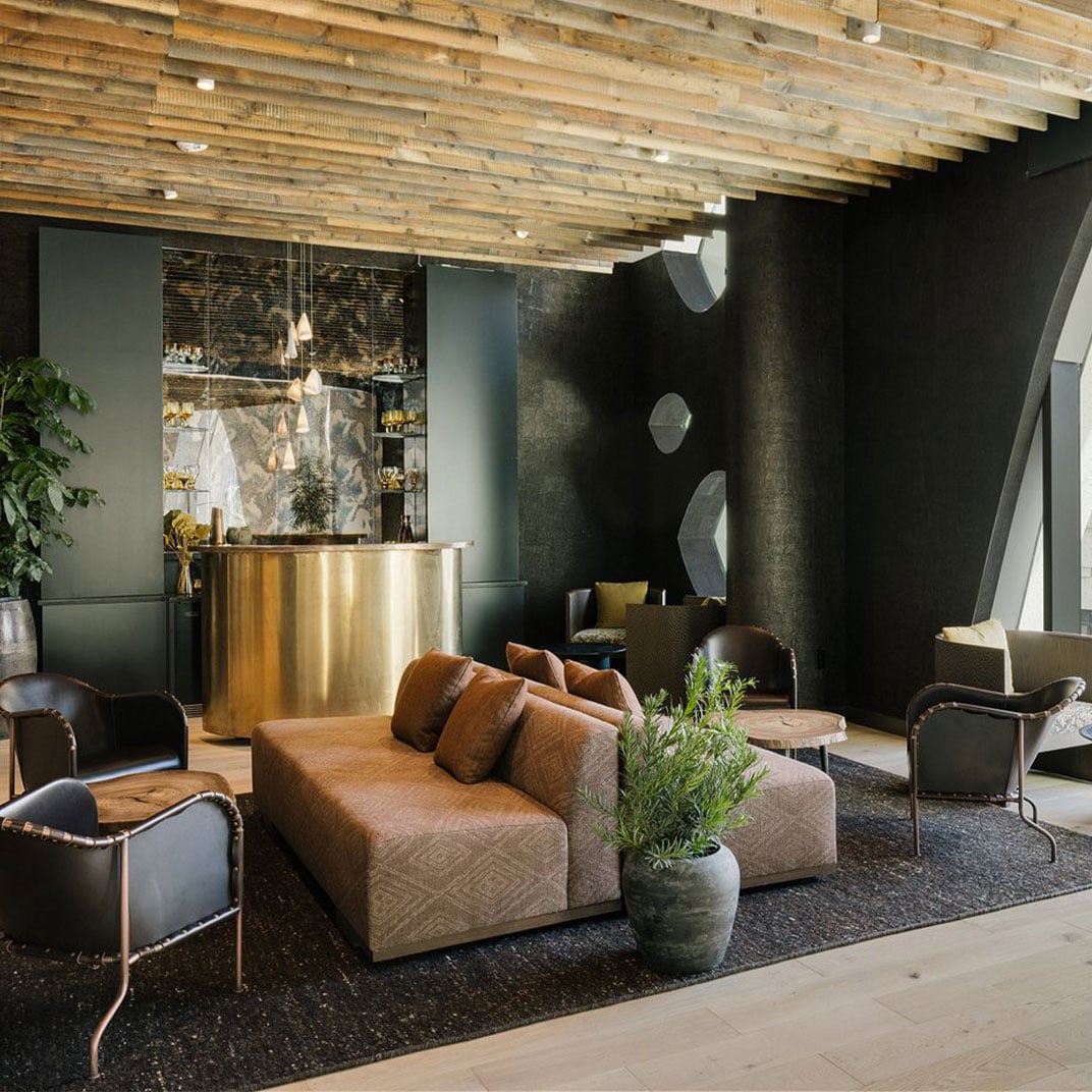 Exposed timber beams crown a sophisticated lobby where rust-colored sectionals and gleaming brass reception desk create an inviting urban retreat beneath dramatic black accent walls.