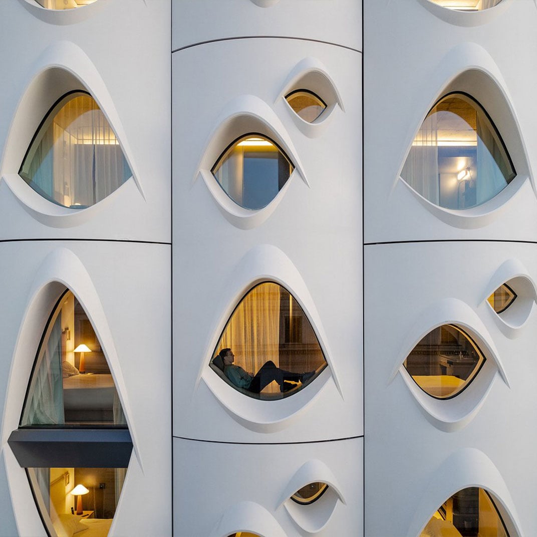 Honeycomb windows reveal intimate capsule-style accommodations where guests curl up in cozy pods, each glowing warmly against the building's sculptural white facade.