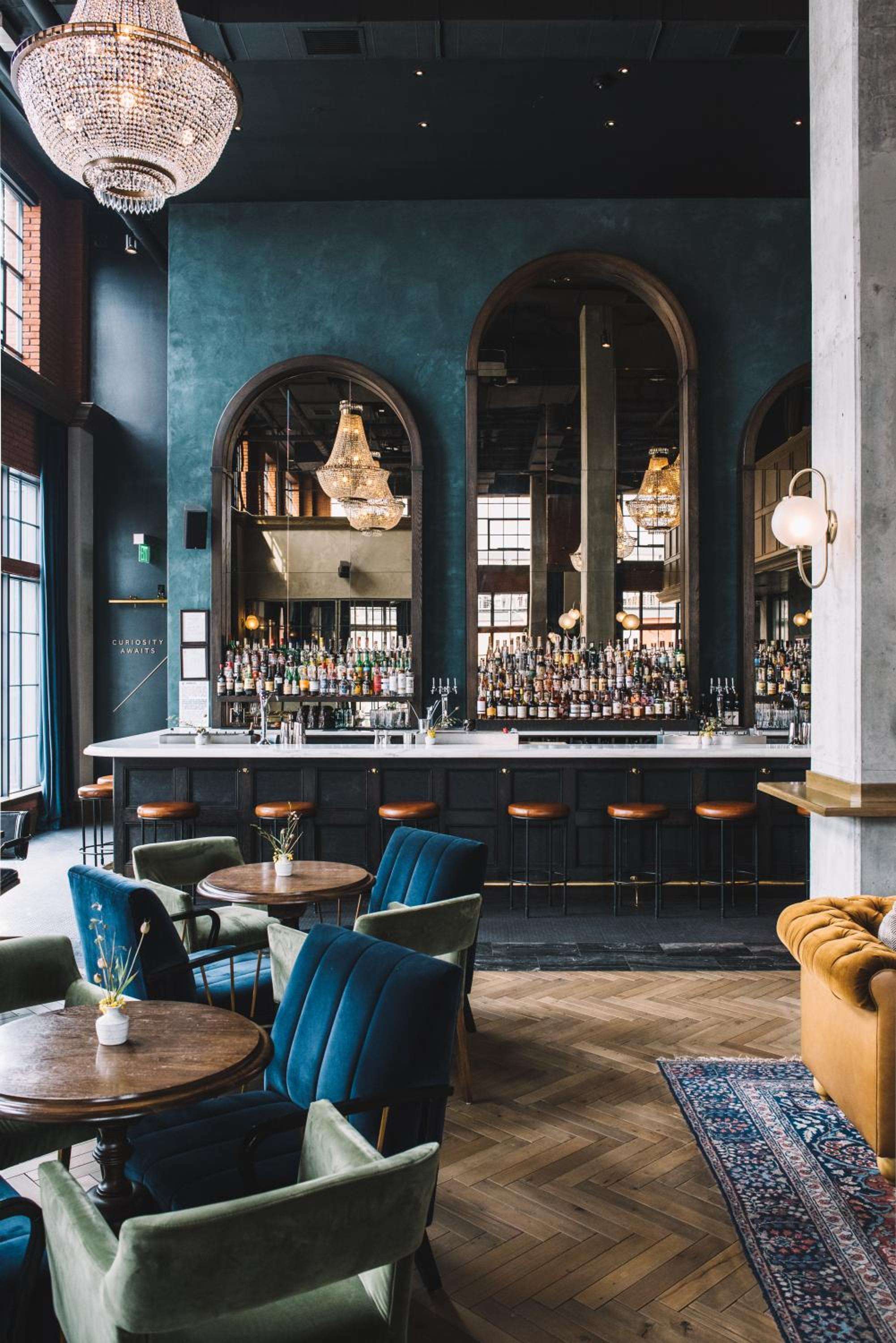 Crystal chandeliers cast warm light over jewel-toned velvet seating and polished brass details, while an impressive spirits collection gleams behind arched mirrors in this sophisticated cocktail haven.