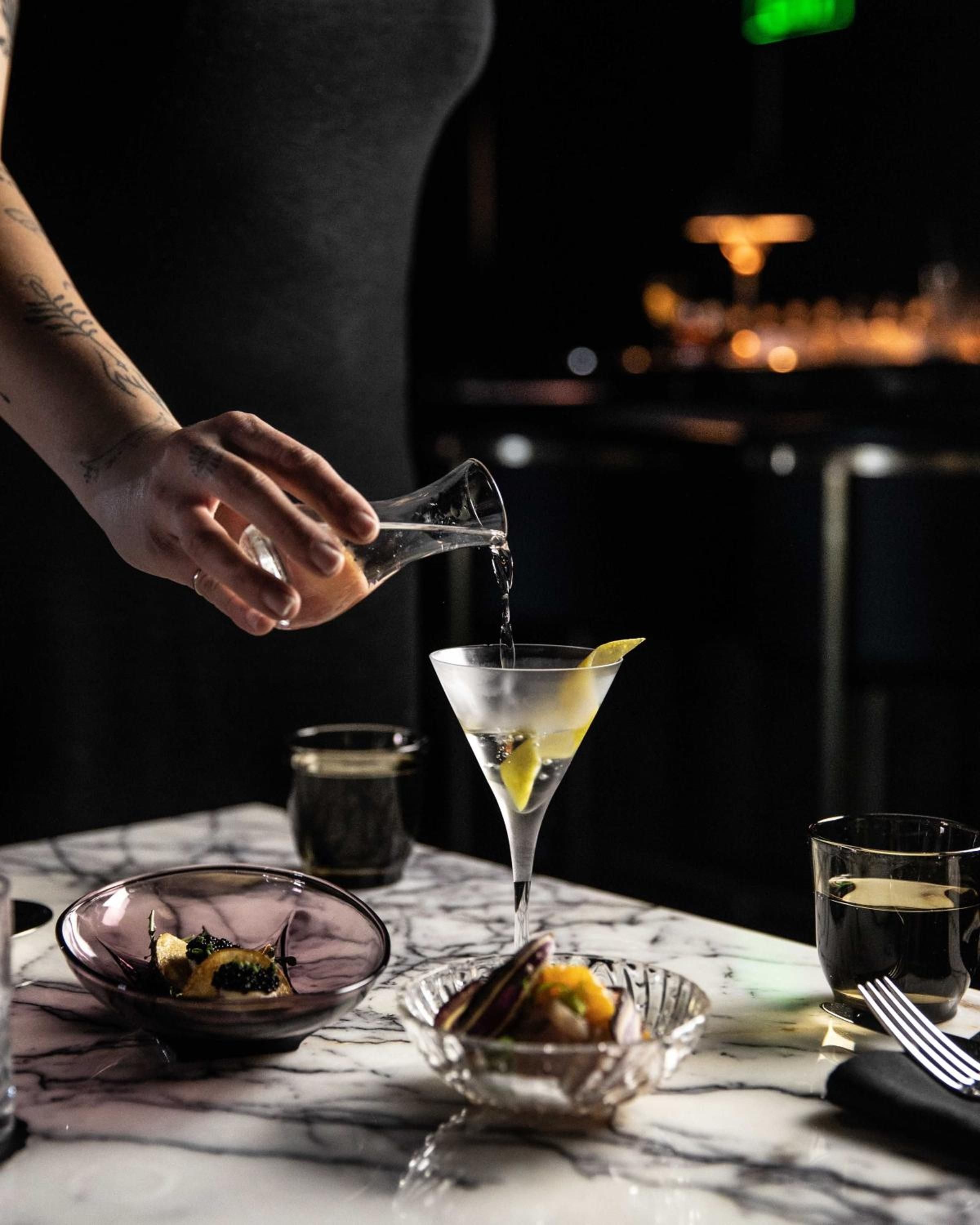 Precise cocktail artistry unfolds as a bartender's tattooed hand pours clear spirits into a perfect martini, accompanied by glistening caviar service on marble.