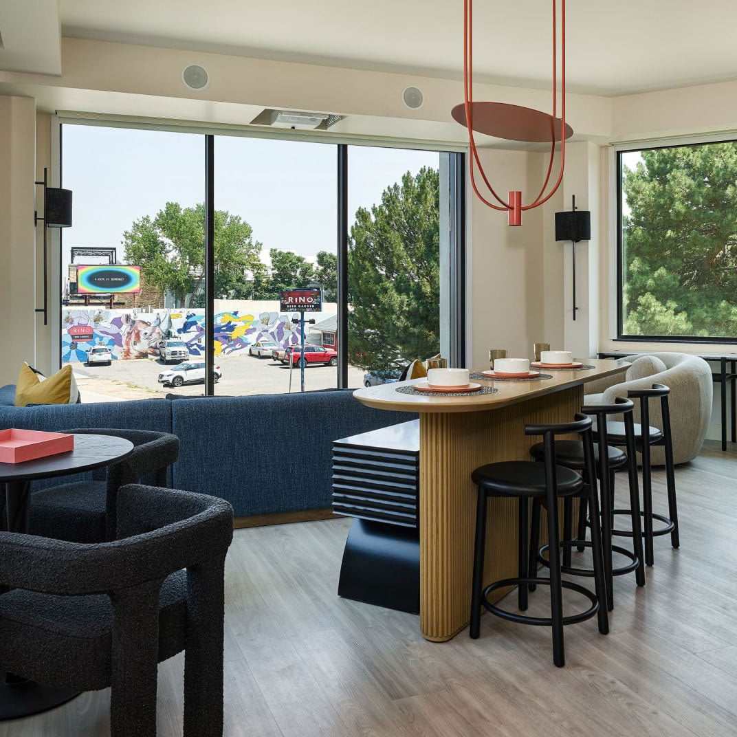 Floor-to-ceiling windows frame colorful street murals while guests gather around the curved bar beneath sculptural red pendant lights in this vibrant social hub.