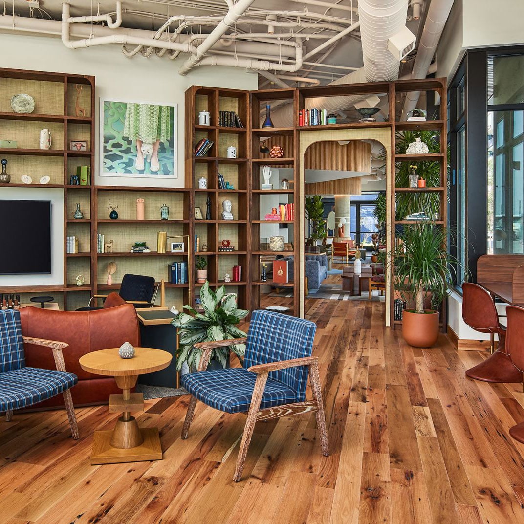 Mid-century charm meets modern comfort in this thoughtfully curated lobby, where blue plaid armchairs and walnut shelving create an inviting literary sanctuary beneath exposed industrial ceilings.
