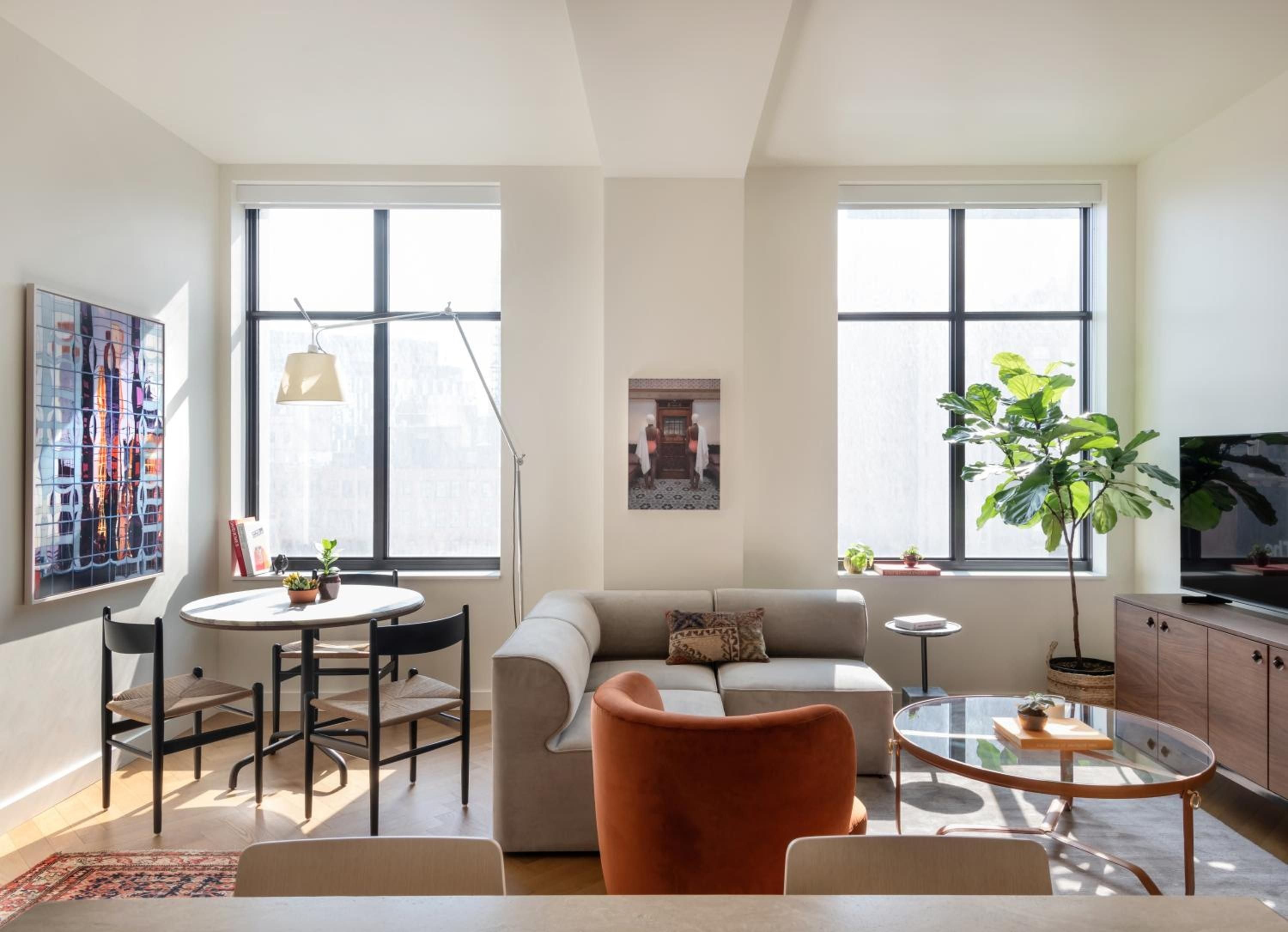 Sunlight streams through oversized windows onto warm wood floors, where mid-century furniture and a vibrant orange chair create an inviting urban sanctuary above Detroit's streets.